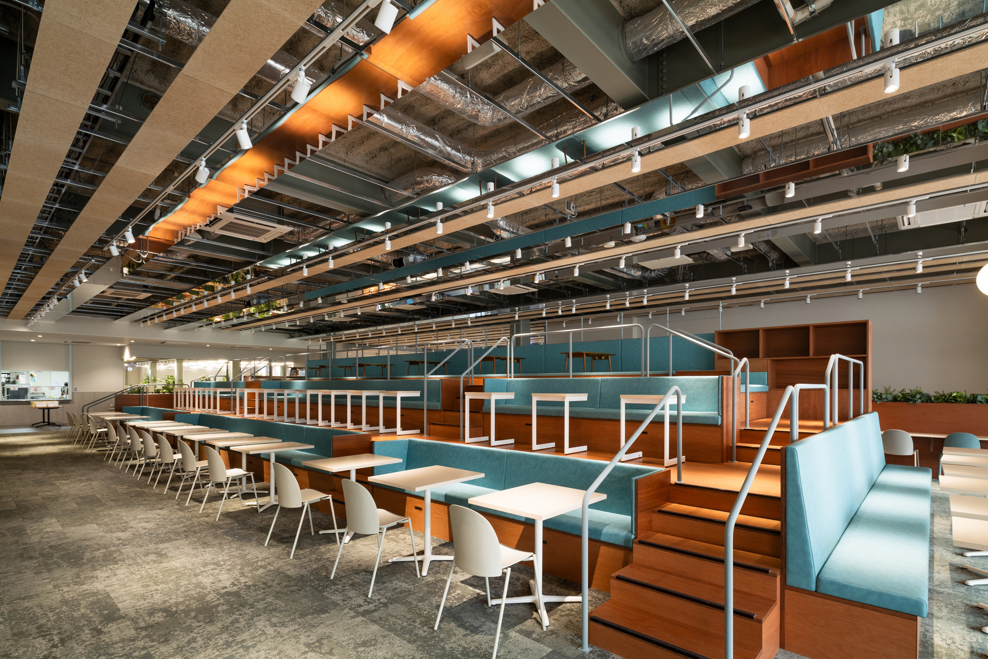 Cafeteria seating with teal upholstered banquettes and exposed ceiling ducts running overhead in parallel lines