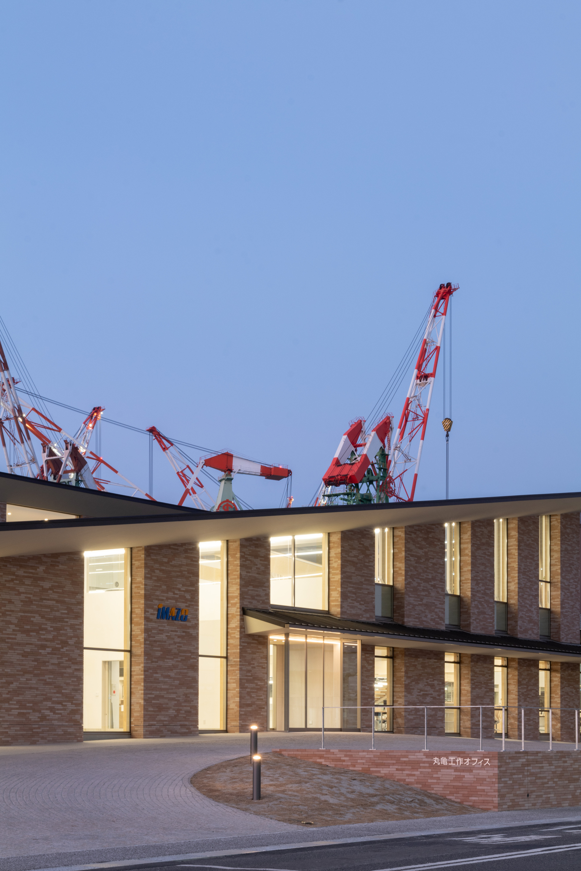Two-story brick facade with floor-to-ceiling glazing illuminated at dusk beside the curved paved forecourt