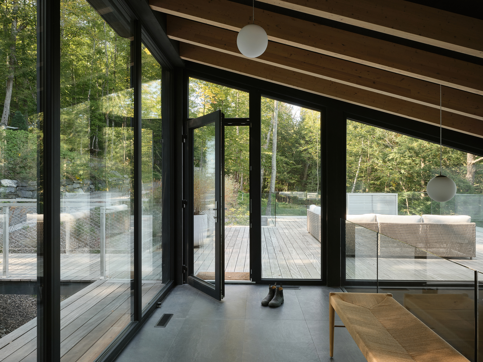 Black-framed folding glass doors opening from concrete floor to timber deck with glass railing in wooded setting