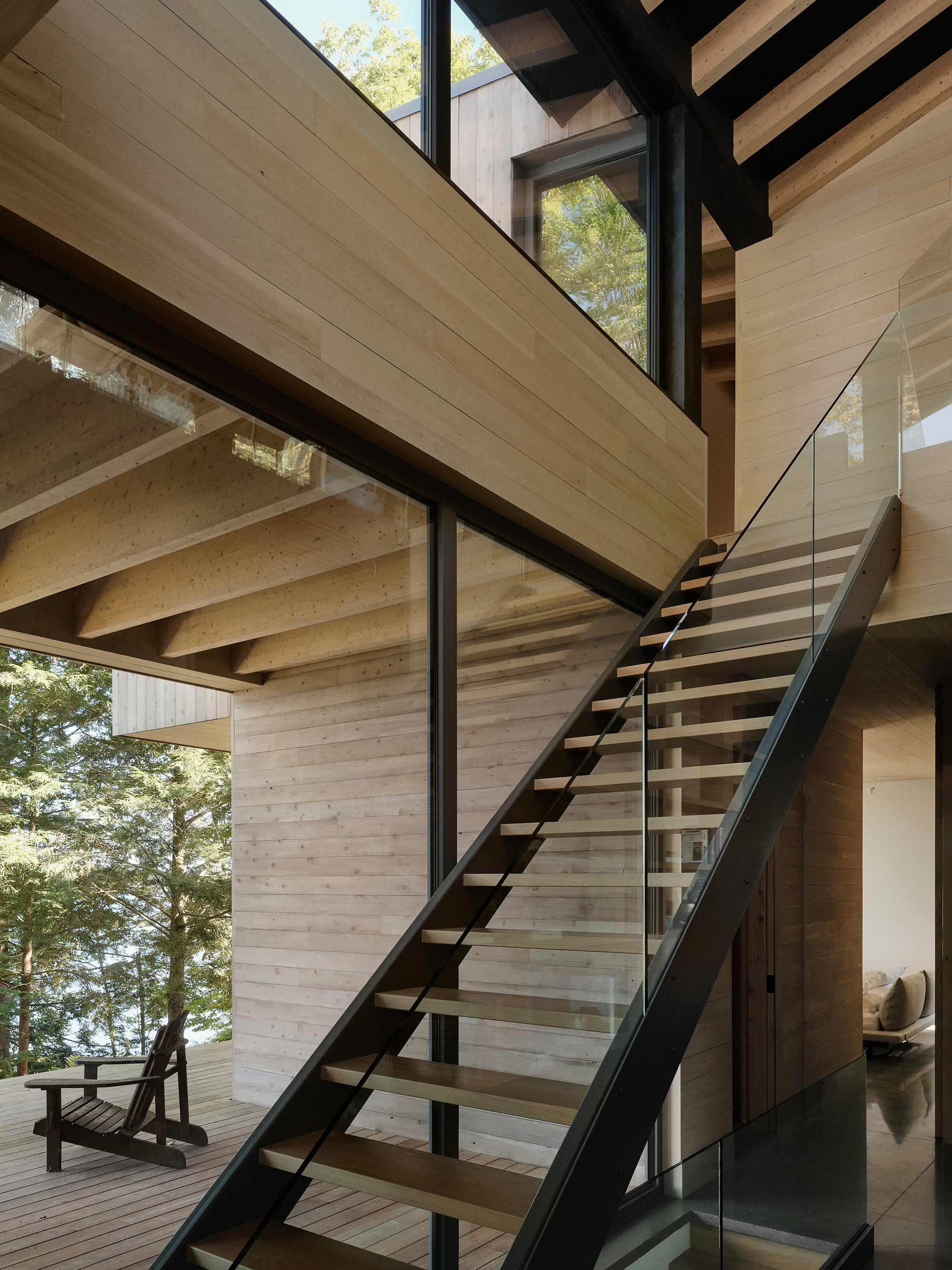Open stair with steel stringers and glass railing ascending through a double-height timber-clad volume overlooking trees