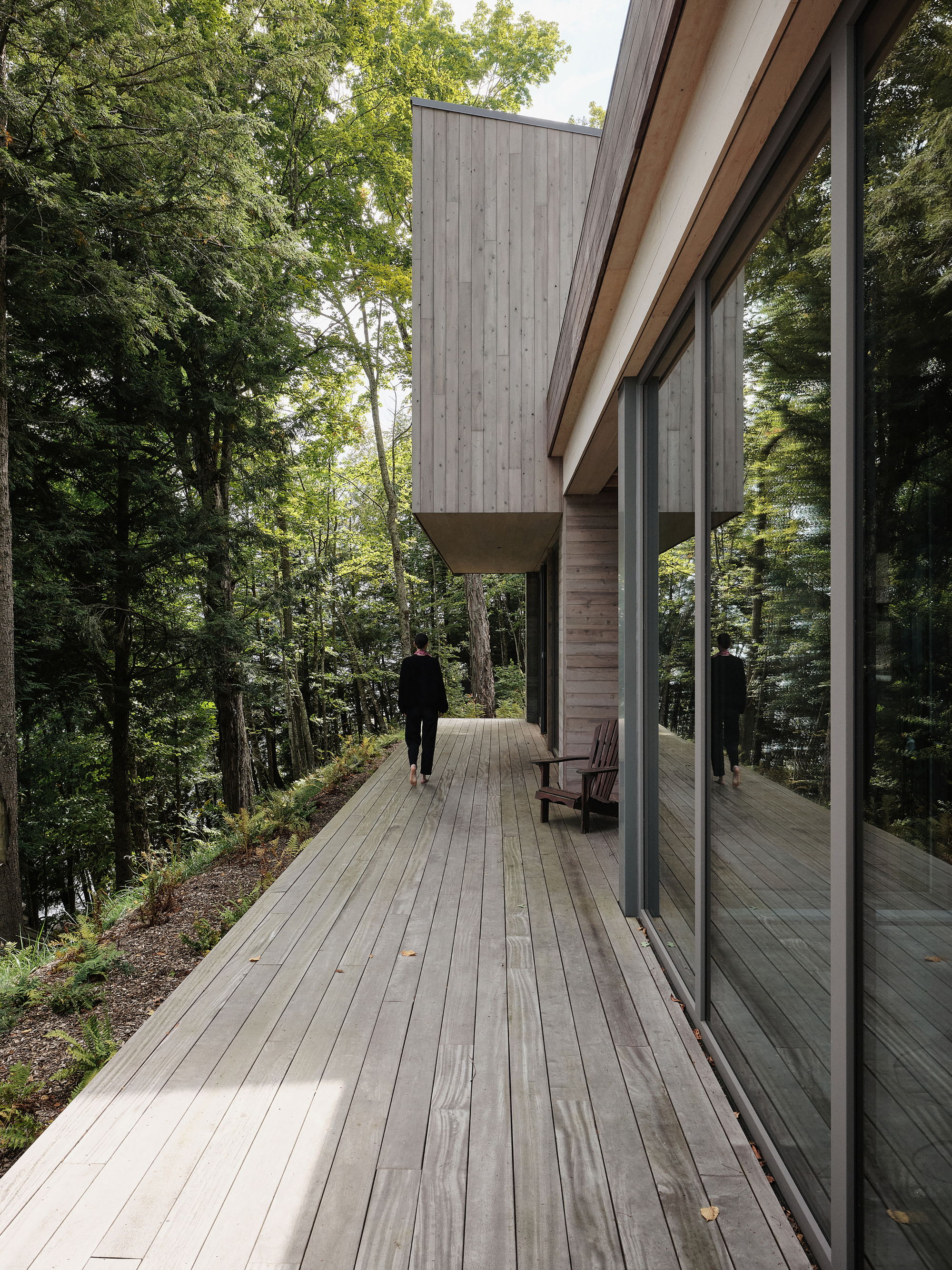 Timber deck running along the glass facade with a person walking past a chair in the forest