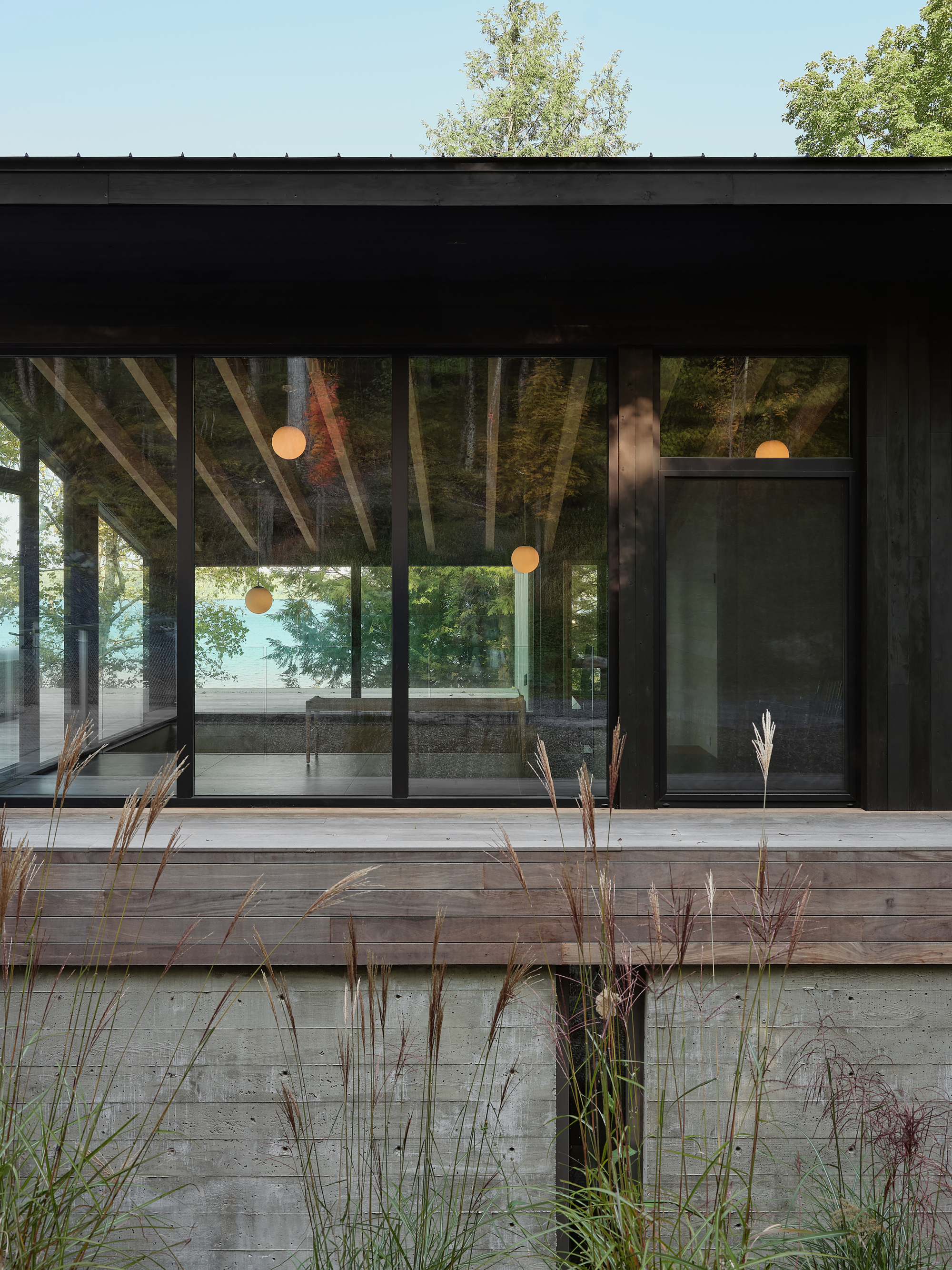Exterior view of glazed facade with pendant lights visible inside and ornamental grasses in foreground at dusk