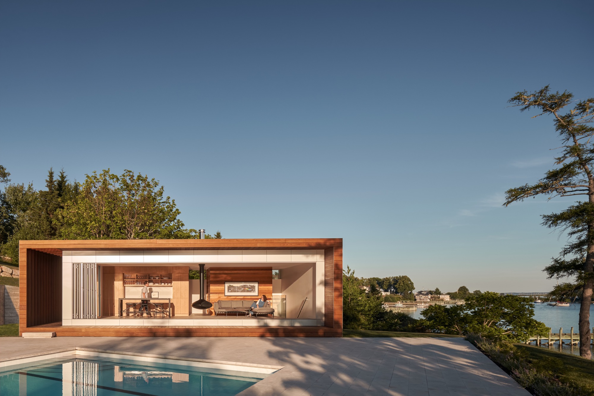 Timber-clad poolside pavilion with open living space overlooking the water at golden hour