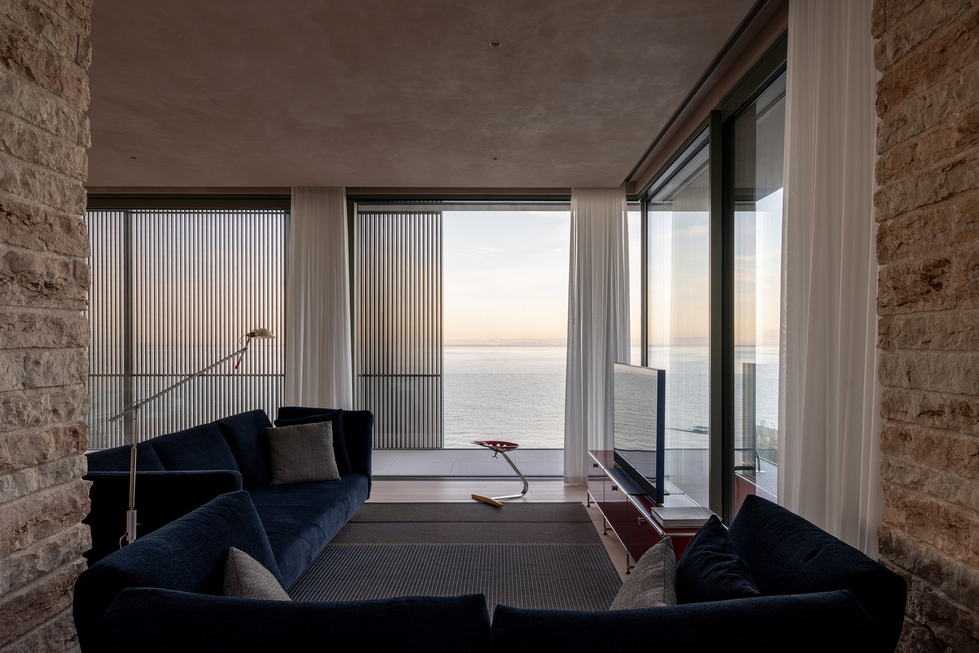 Living room with floor-to-ceiling glazing, rammed earth walls and water views at twilight