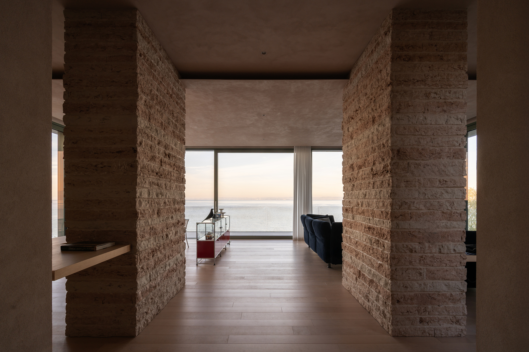Interior hallway framed by rough stone columns leading to a glazed opening with ocean views at dusk