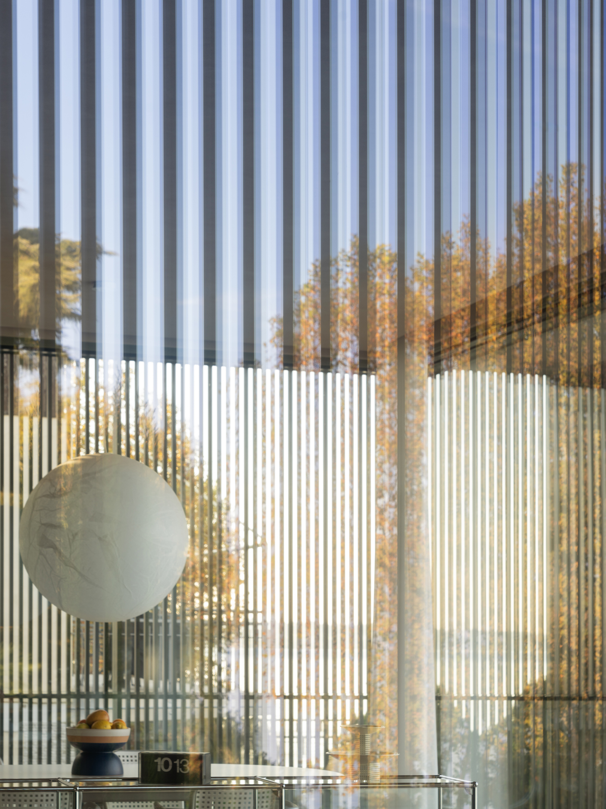 Vertical metal louvers filtering daylight onto spherical pendant above mirrored dining shelf with autumn trees beyond
