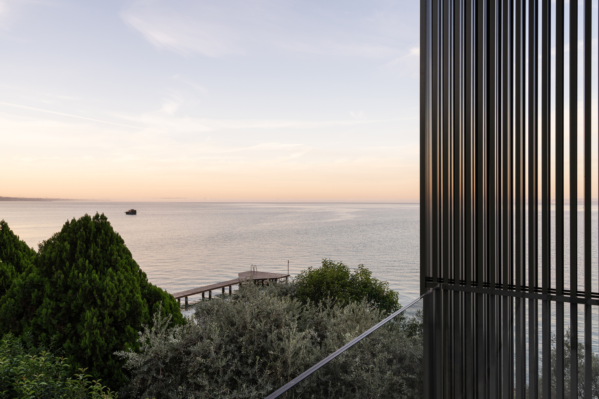 Corner of dark vertical screen facade overlooking calm water, planted slope and timber jetty at sunset