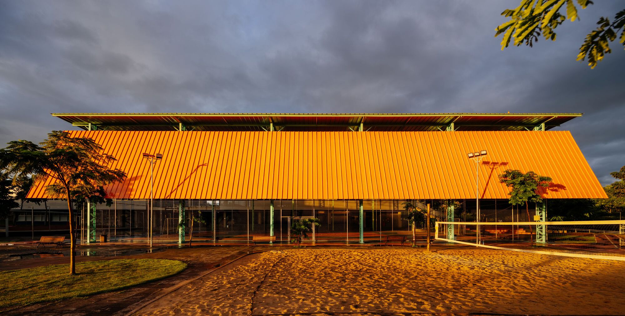Glazed pavilion with an orange corrugated metal roof illuminated by golden hour sunlight under stormy clouds