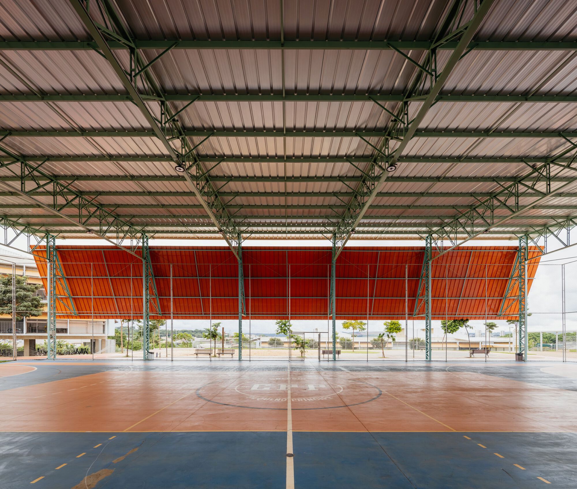 Covered sports court with green steel trusses and orange perforated wall panels behind planted trees