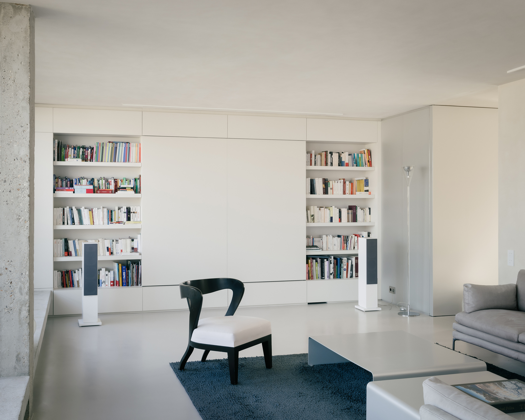 White storage wall with recessed bookshelves flanking a central panel beside a concrete column
