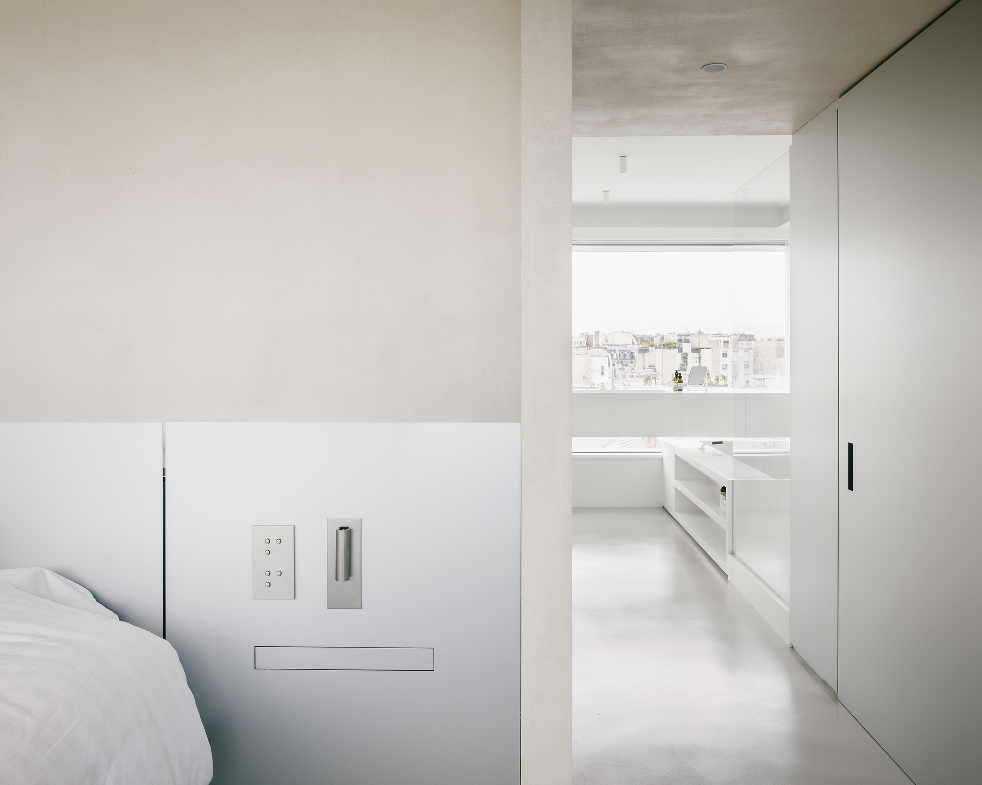 View through bedroom doorway into bathroom with window framing distant rooftops