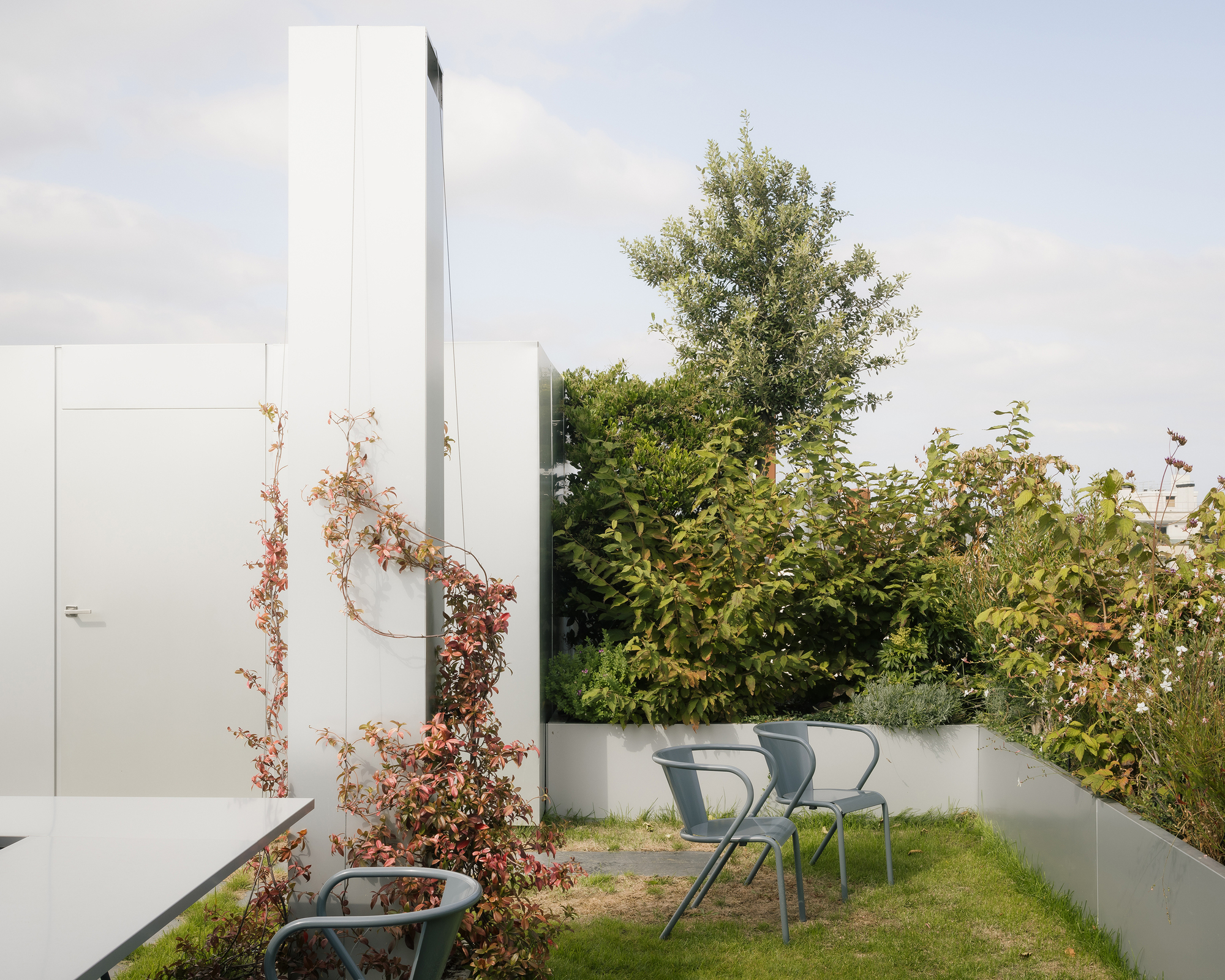Rooftop terrace with white metal chairs and climbing vines along white walls beneath a tall chimney