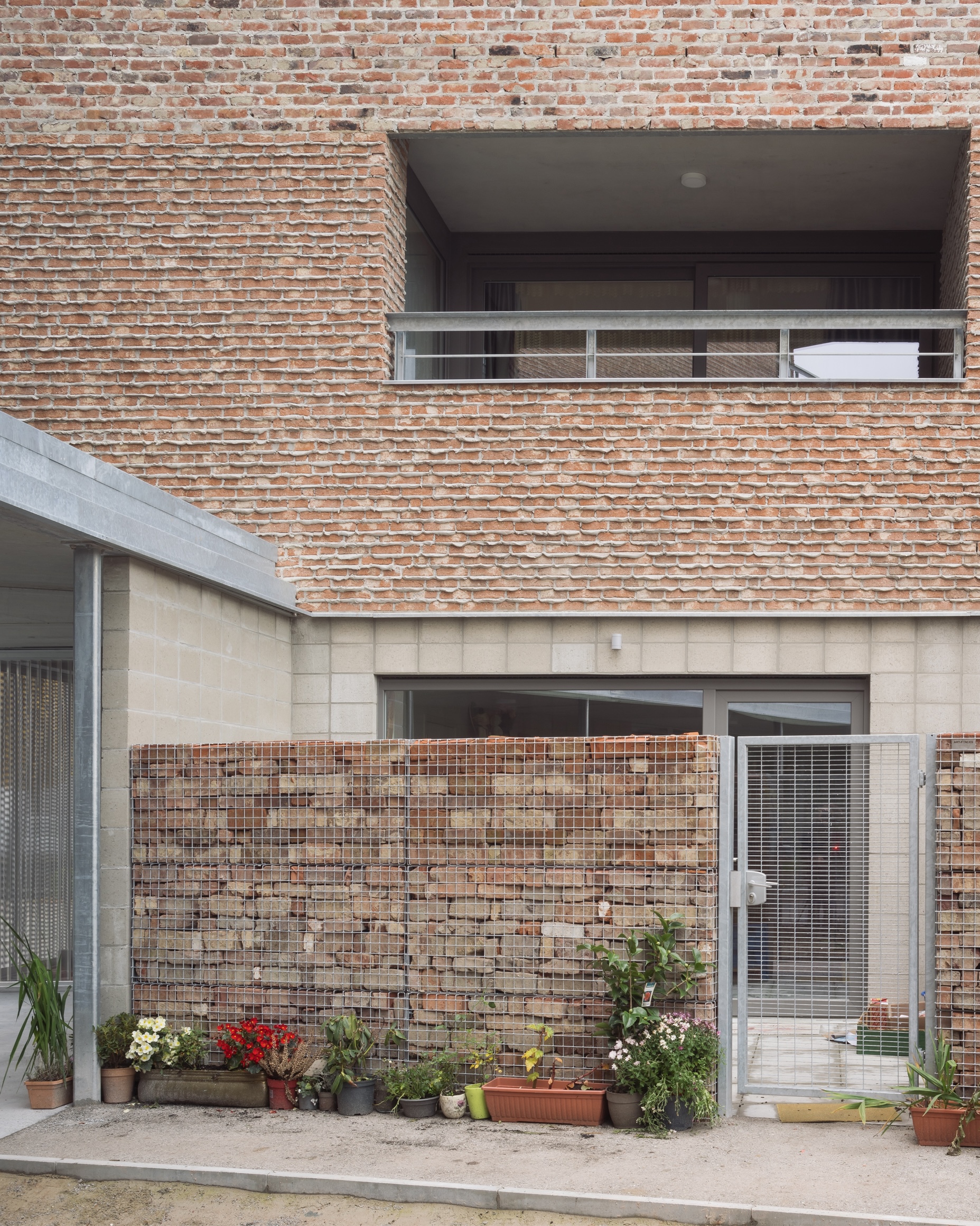 Ground floor courtyard with gabion wall and potted plants beneath cantilevered brick facade and balcony