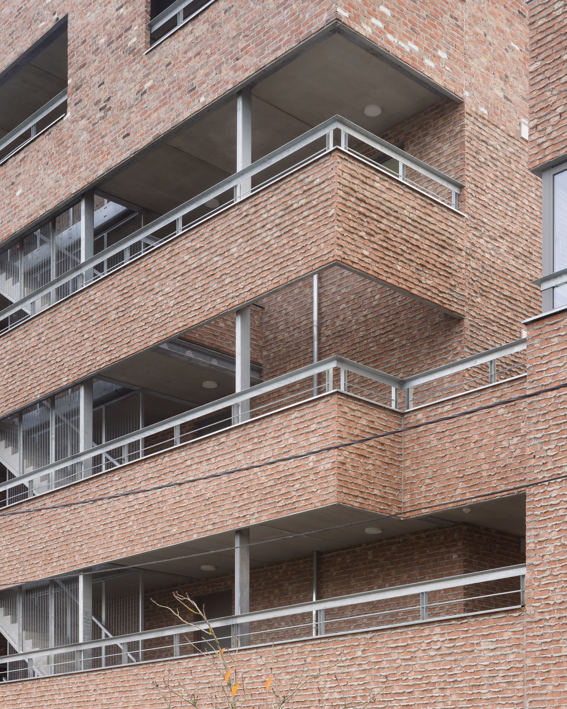 Facade detail of stacked brick balconies with recessed terraces and aluminum handrails under overcast skies