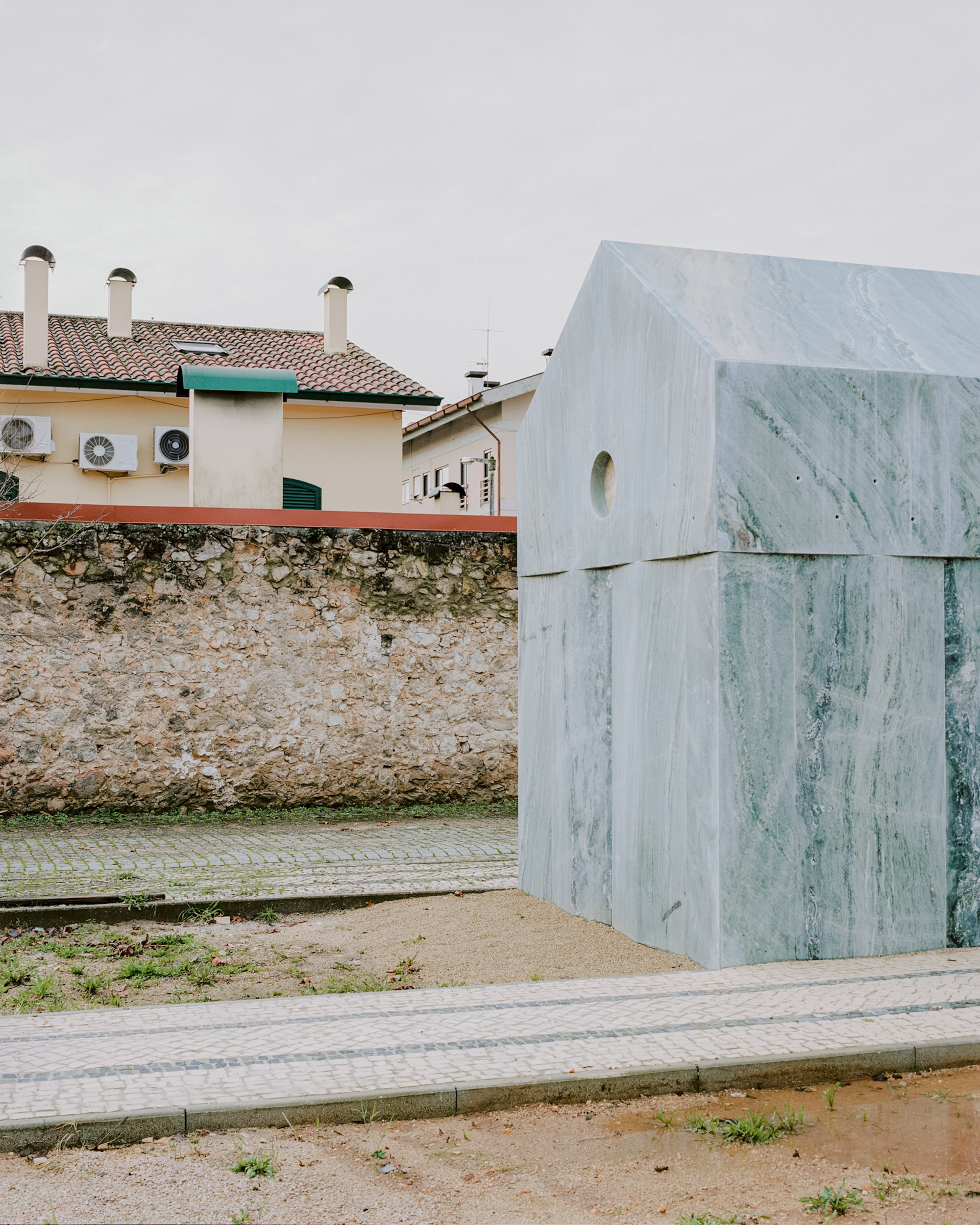 Side elevation showing layered marble panels against a weathered stone boundary wall and neighboring houses