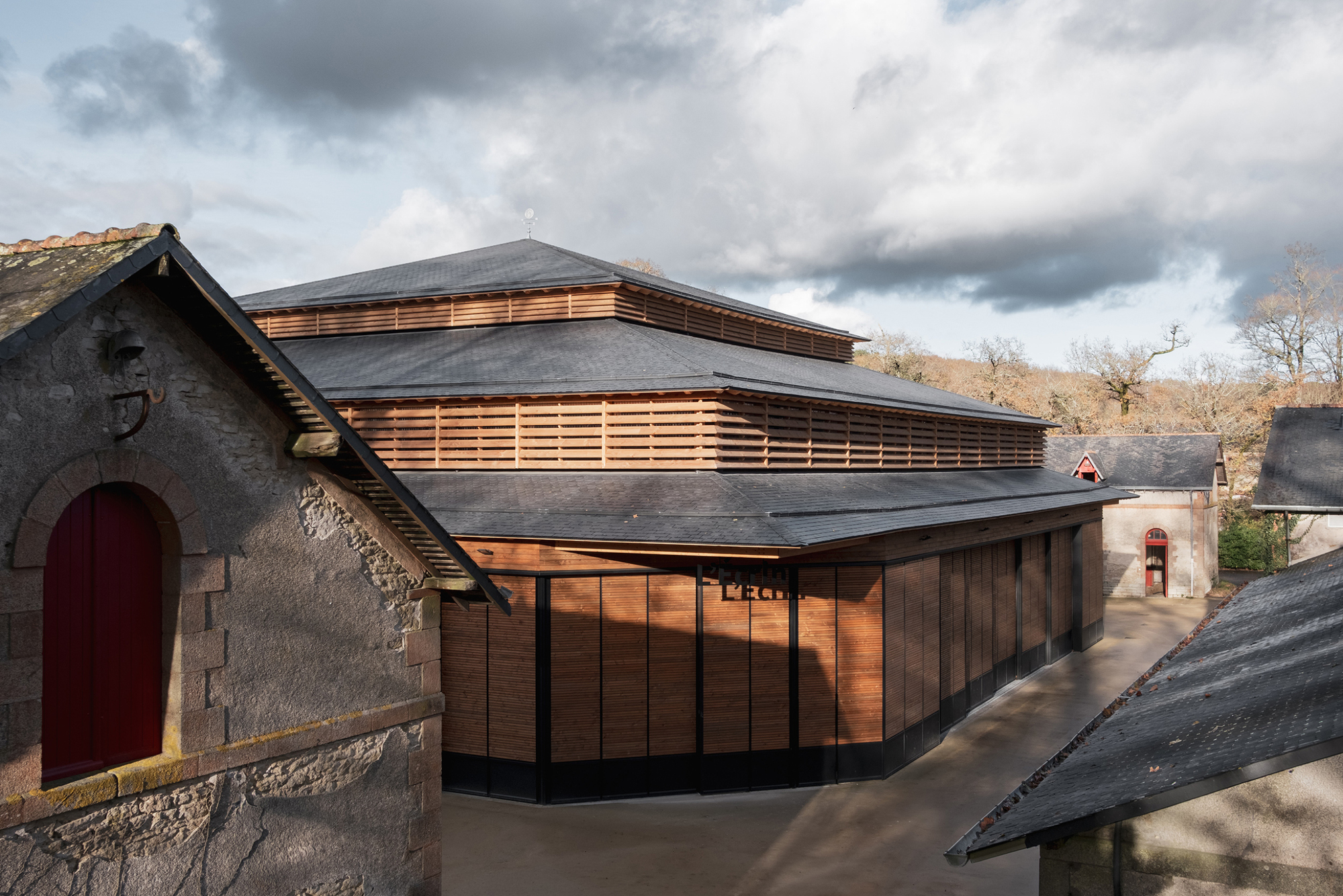 Elevated view of the timber-clad volume with horizontal slat screens and multi-level slate roof under dramatic clouds