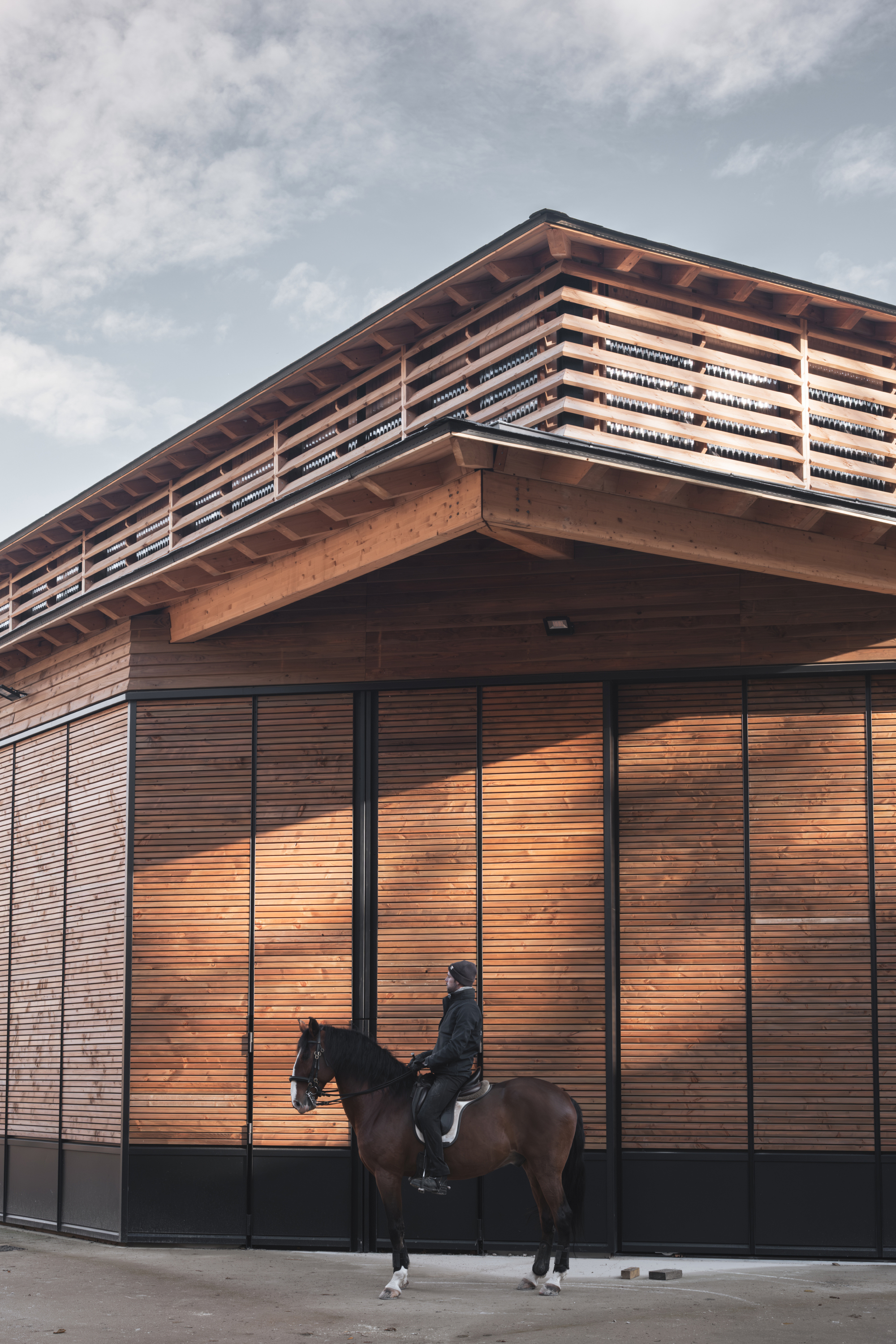 Corner facade of horizontal timber slats with exposed roof rafters and a rider on horseback below
