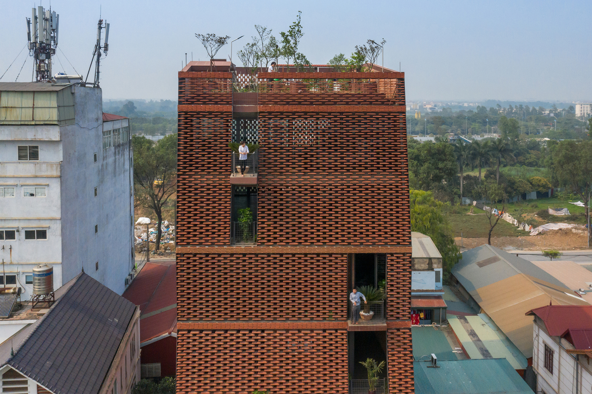 Perforated brick tower rising above neighboring rooftops with planted terraces and people visible in openings