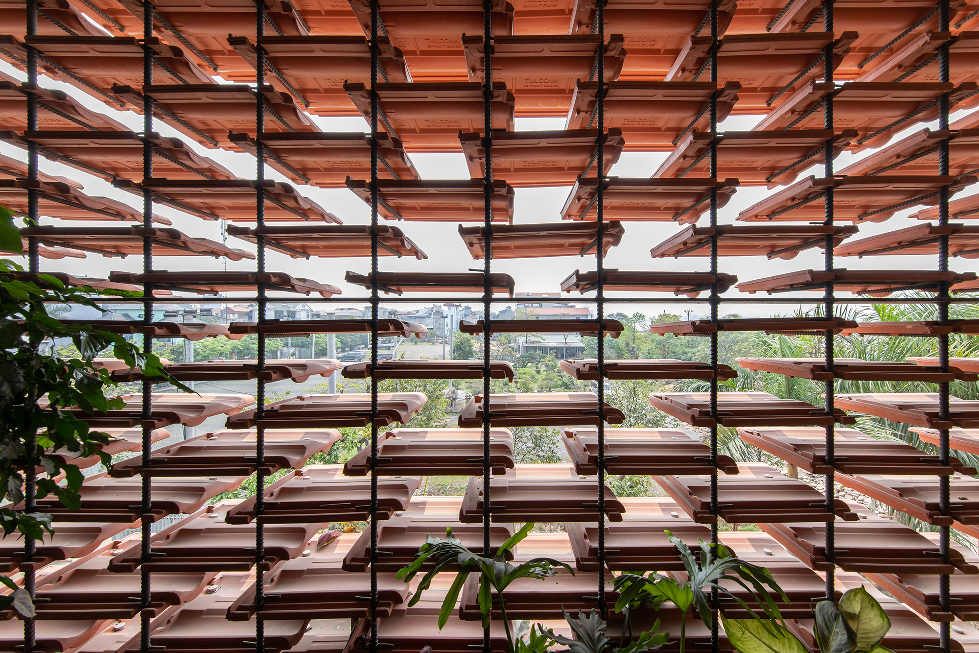Clay tile brise-soleil screen with steel rod supports framing a view of rooftops and greenery