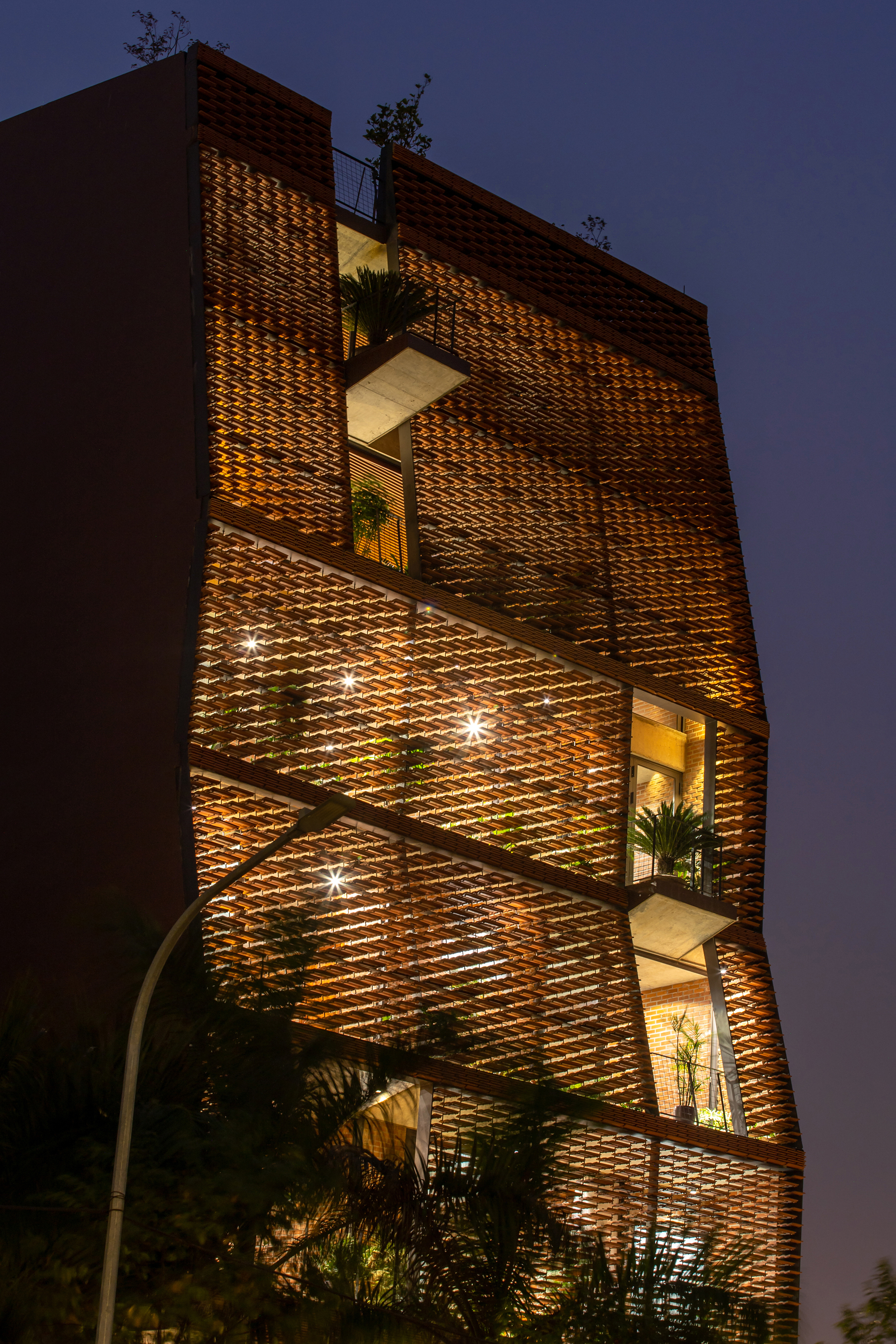 Perforated brick facade with recessed terraces illuminated at dusk and surrounded by trees