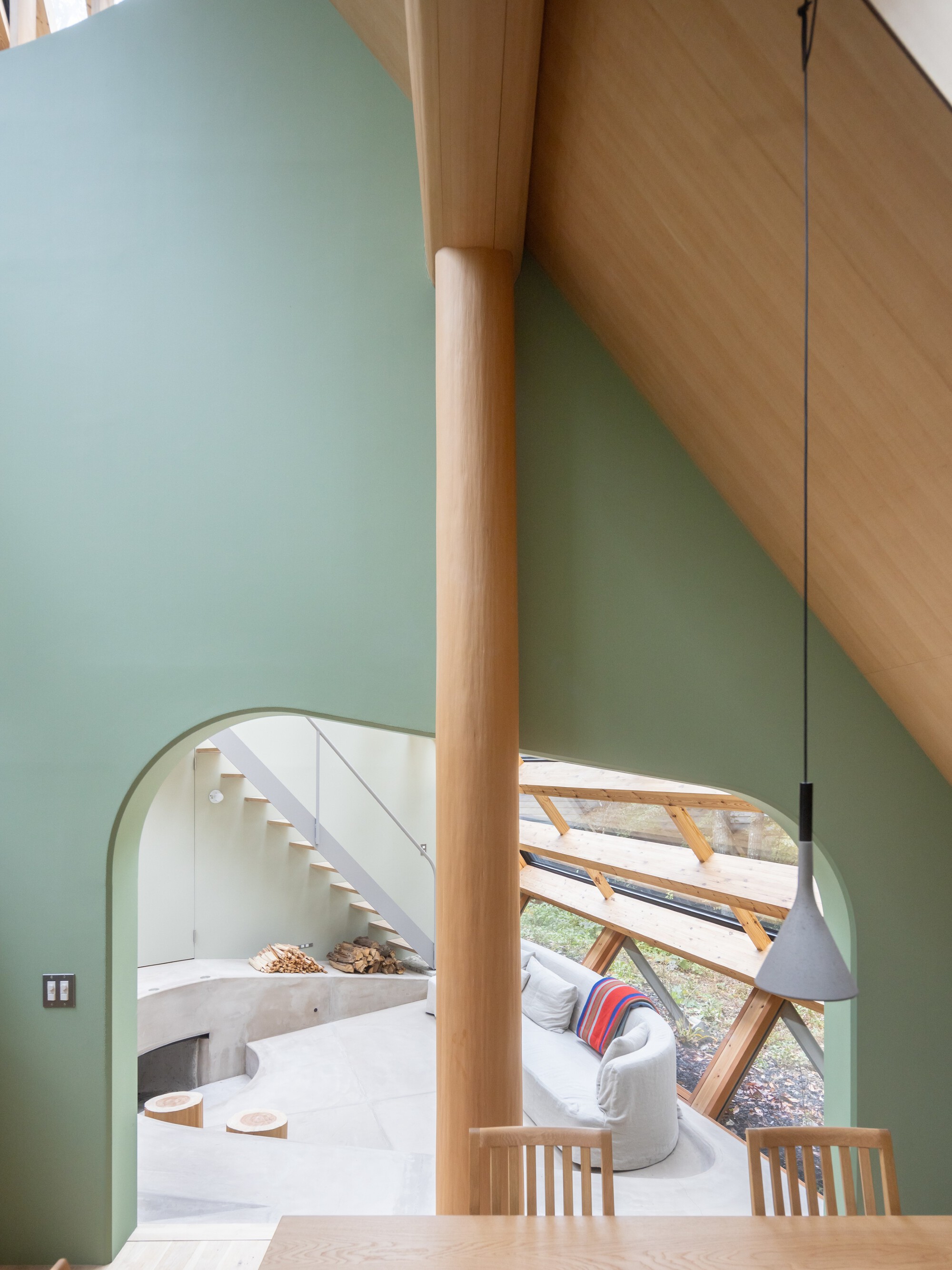 Round timber column between pale green walls with arched opening revealing white stairs and glass-walled living area beyond