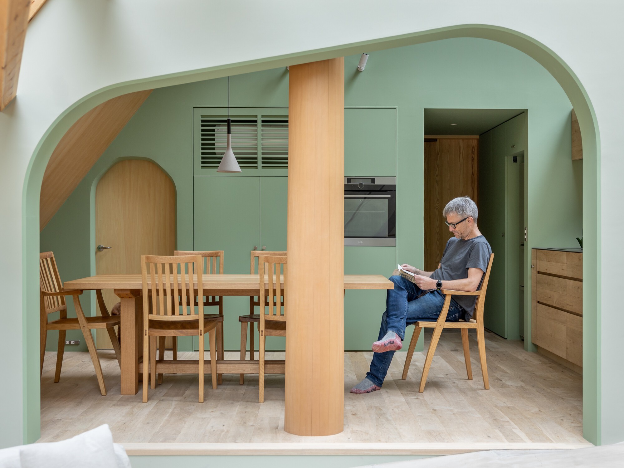 Dining area framed by pale green arched opening with resident reading at timber table beside kitchen cabinets