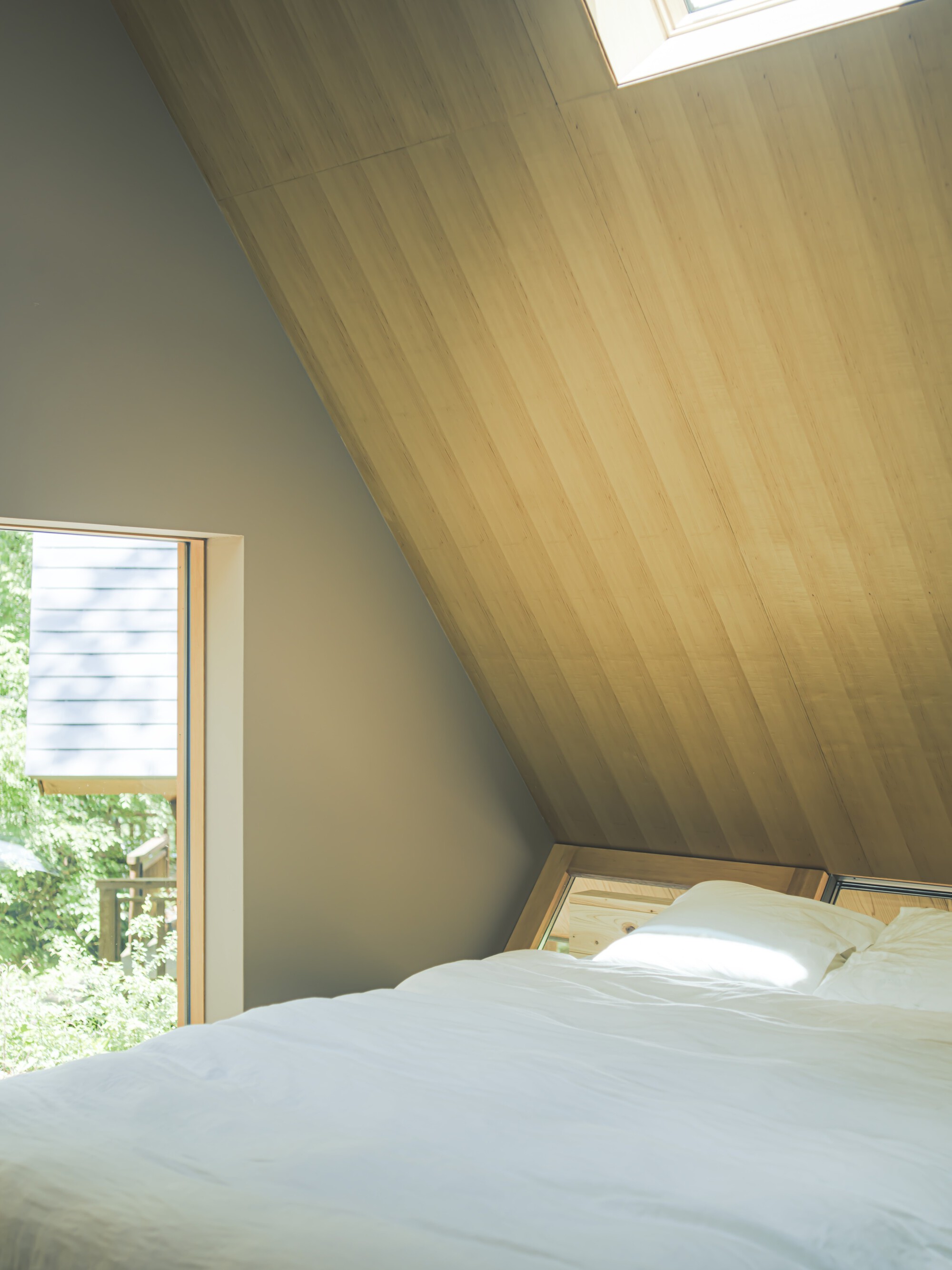 Bedroom with sloped plywood ceiling, corner window, and white bedding in morning light