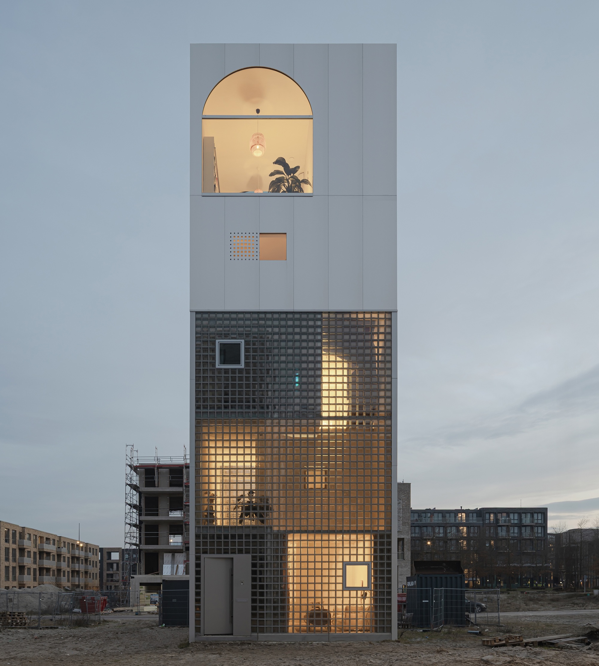 Narrow tower facade with dark glass brick base and pale upper volume framed by an arched window at dusk