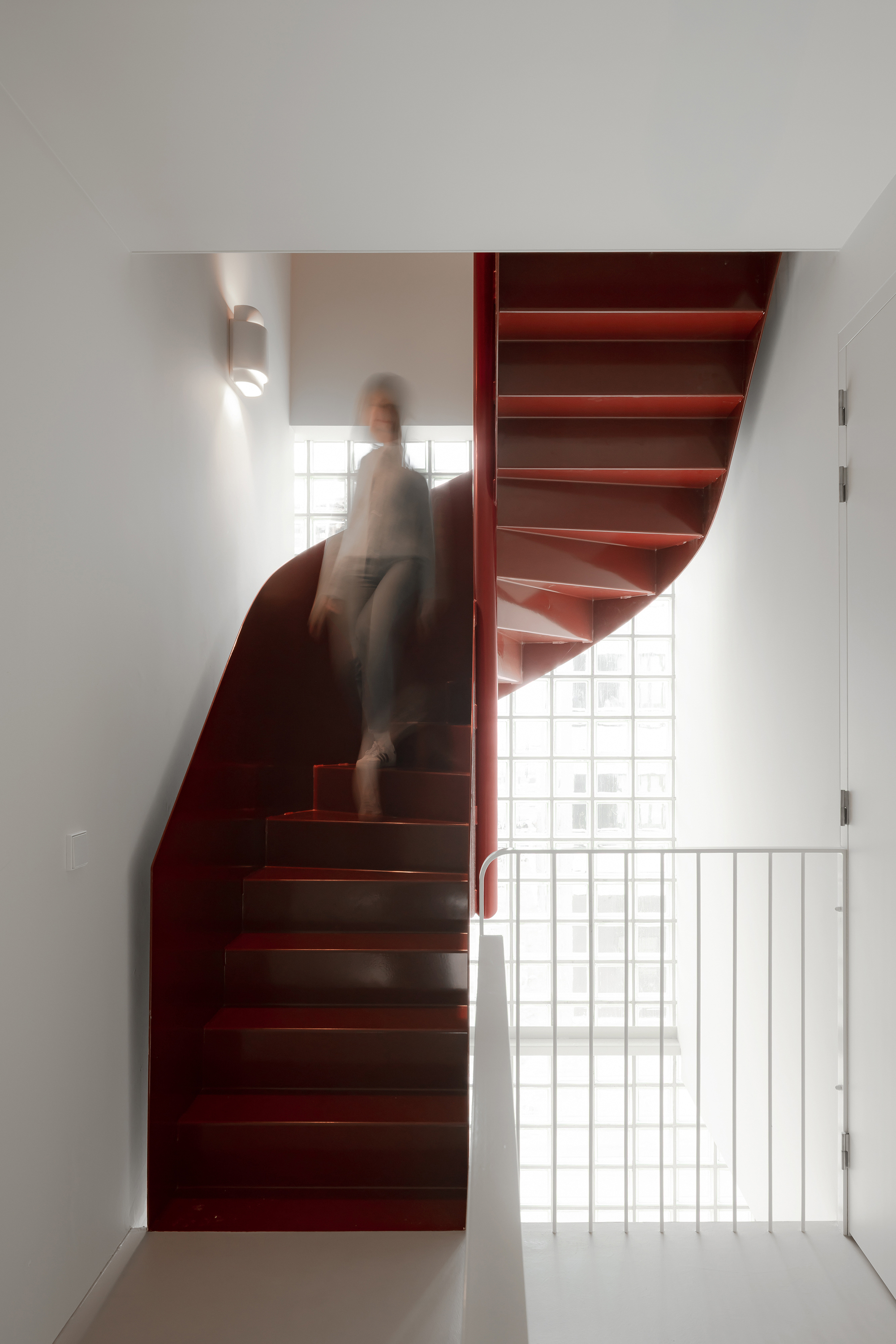 Red steel staircase curving upward with a blurred figure ascending past glass block windows