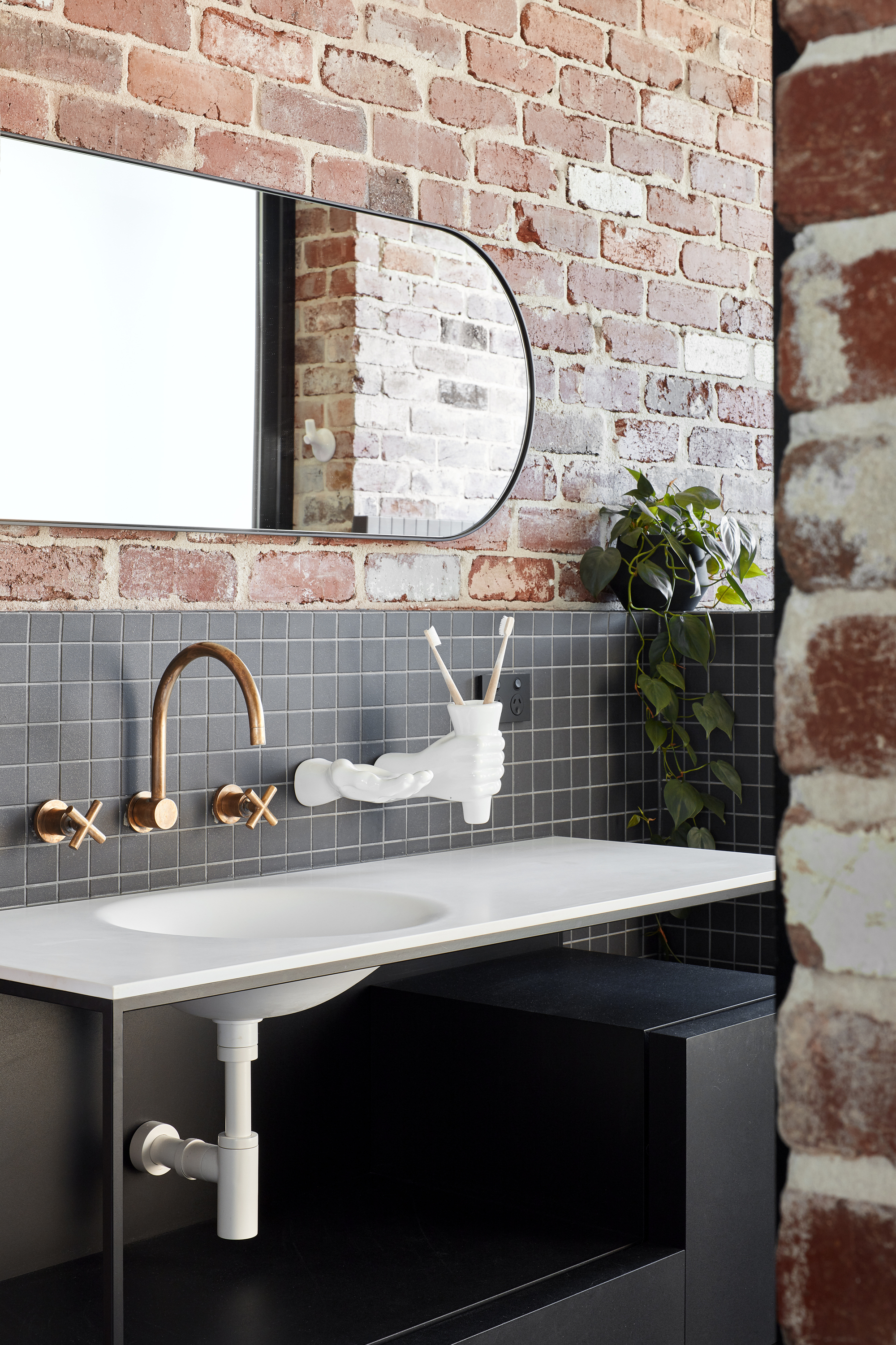 Bathroom vanity with grey mosaic tile backsplash and wall-mounted brass fixtures below exposed brick