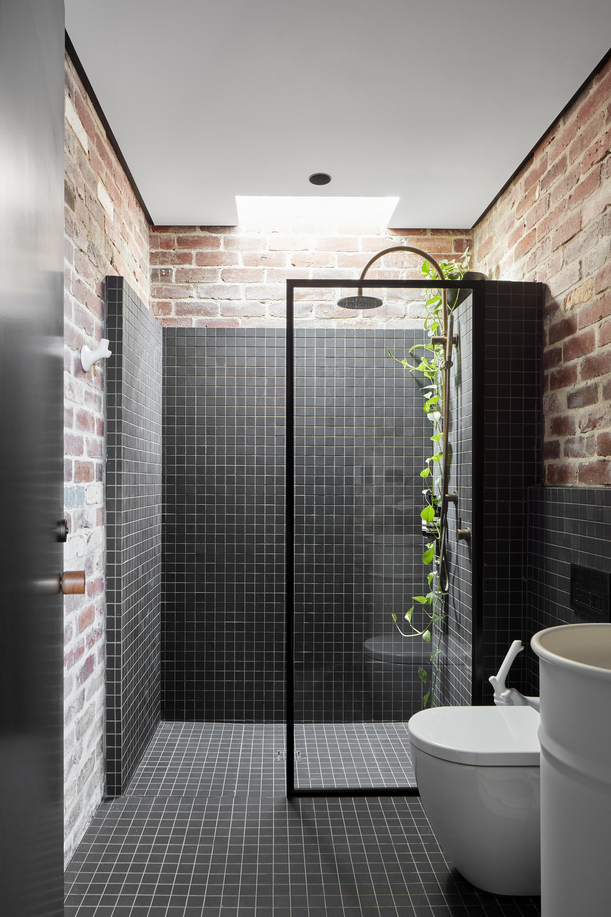 Bathroom with black mosaic tile shower enclosure beneath a skylight between exposed brick walls