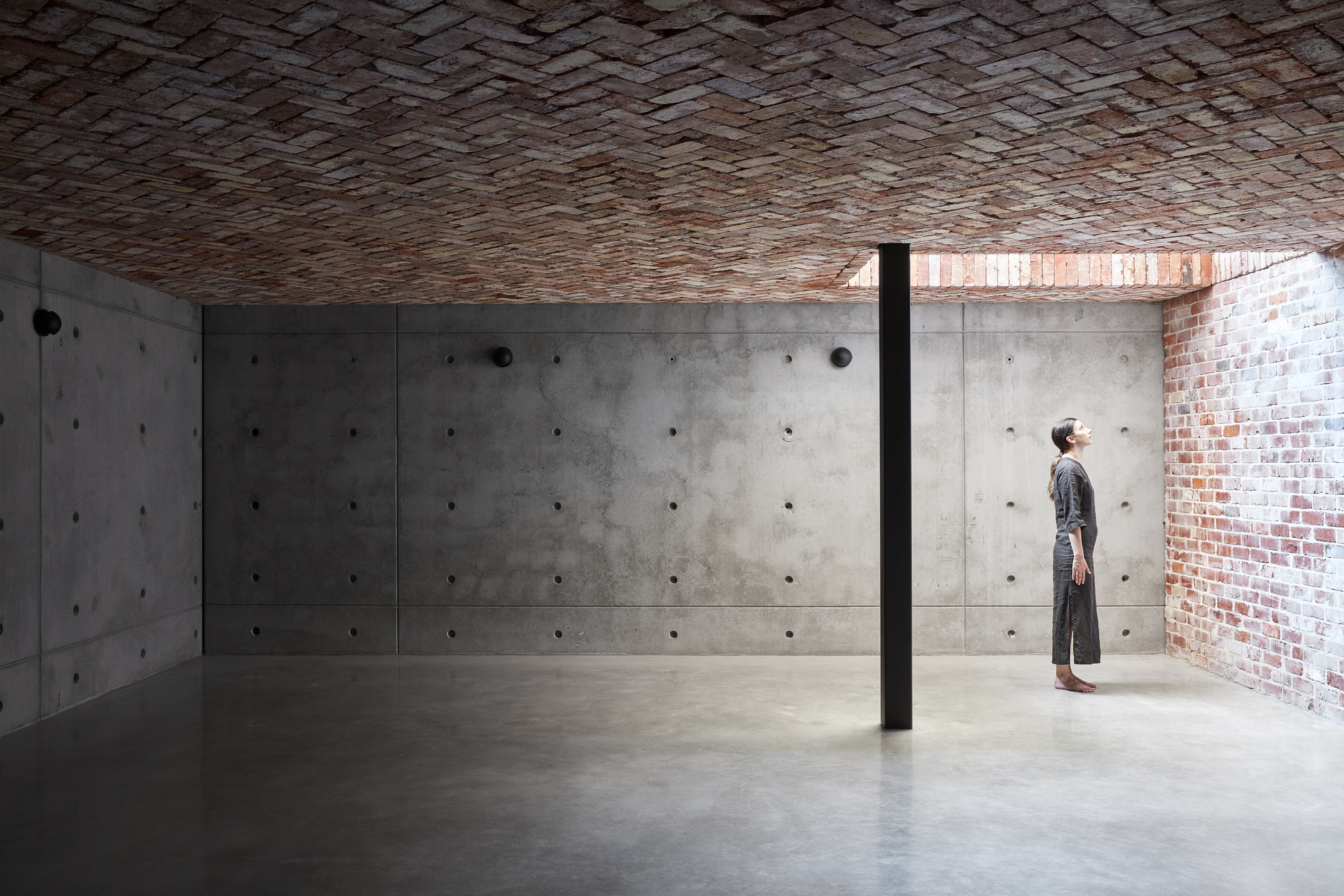 Interior space with board-formed concrete walls beneath a herringbone brick vault ceiling and a single figure
