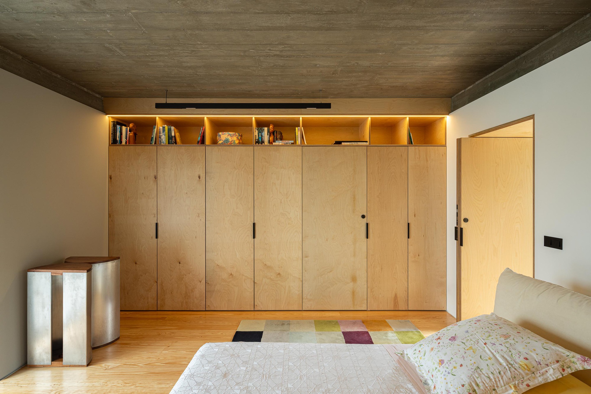 Bedroom with full-height plywood storage wall and backlit upper shelving beneath a timber ceiling