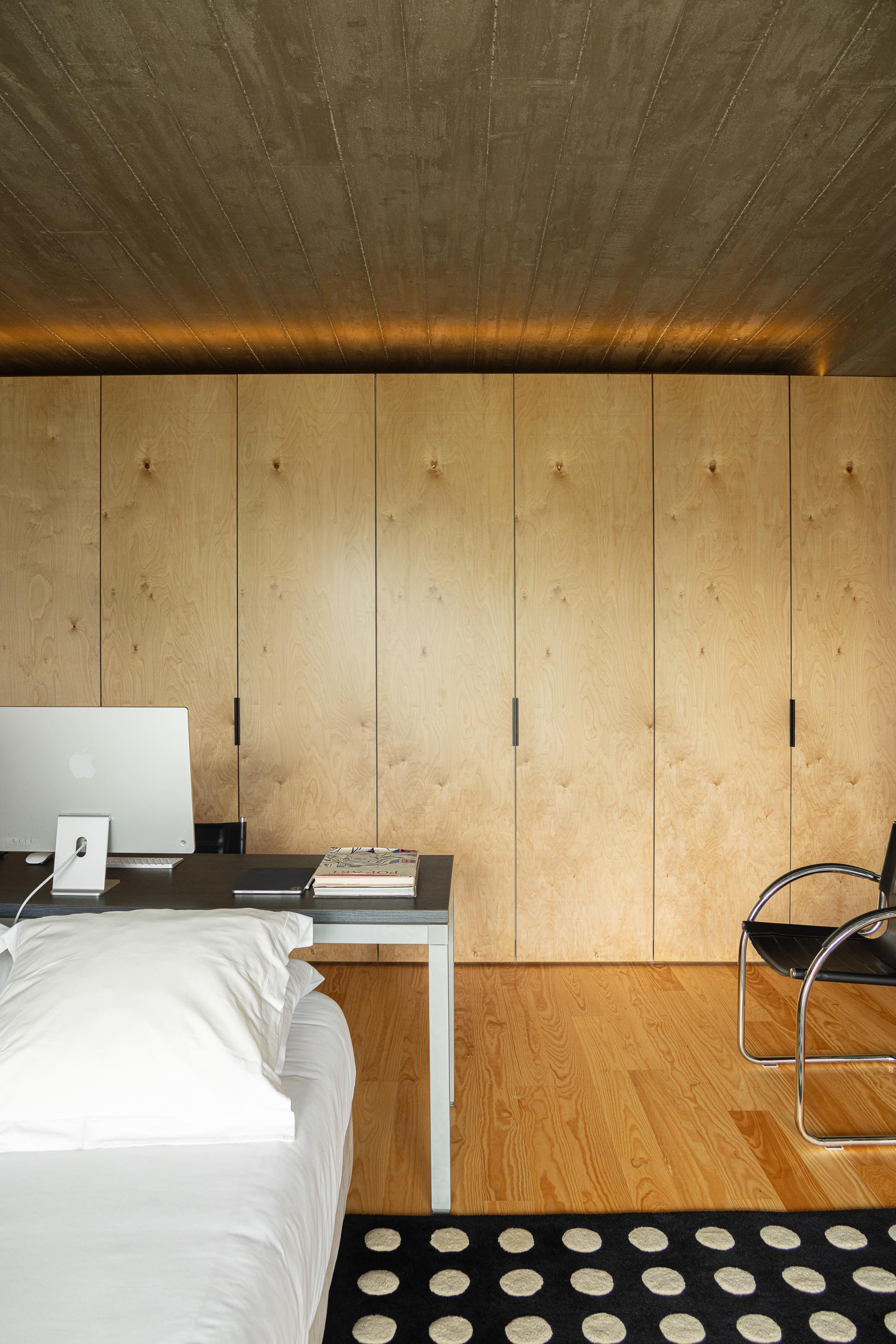 Bedroom with light plywood storage wall, concrete ceiling, and white bedding under warm indirect lighting