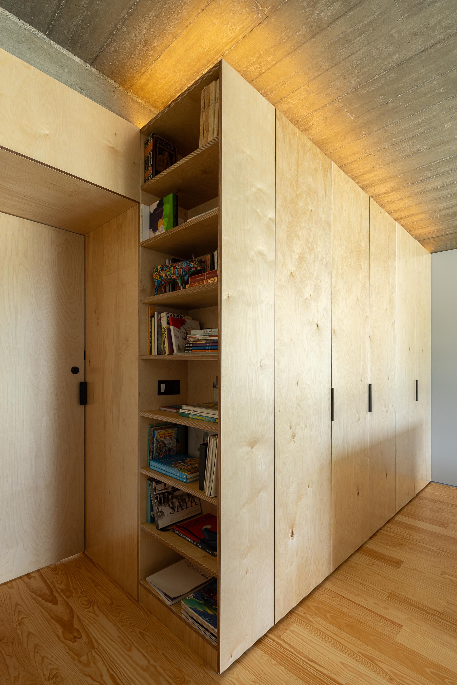 Open plywood cabinet revealing shelves with books beneath a board-formed concrete ceiling