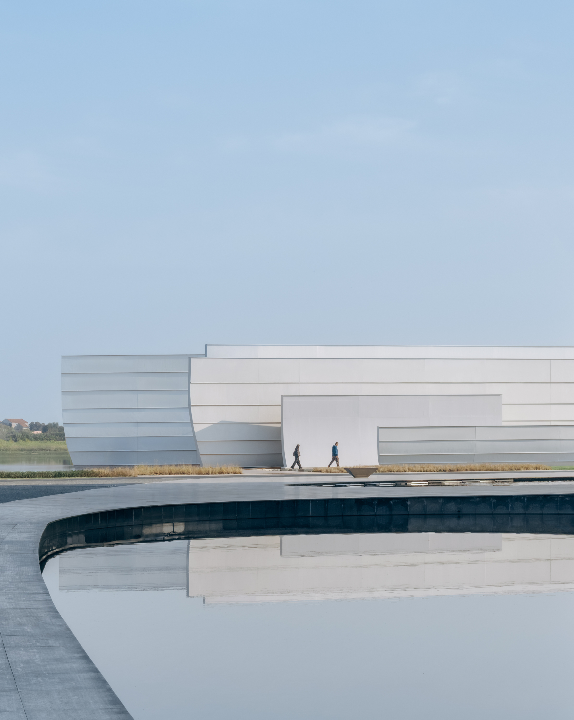 Curved water channel beside the white layered facade with two figures walking on the plaza