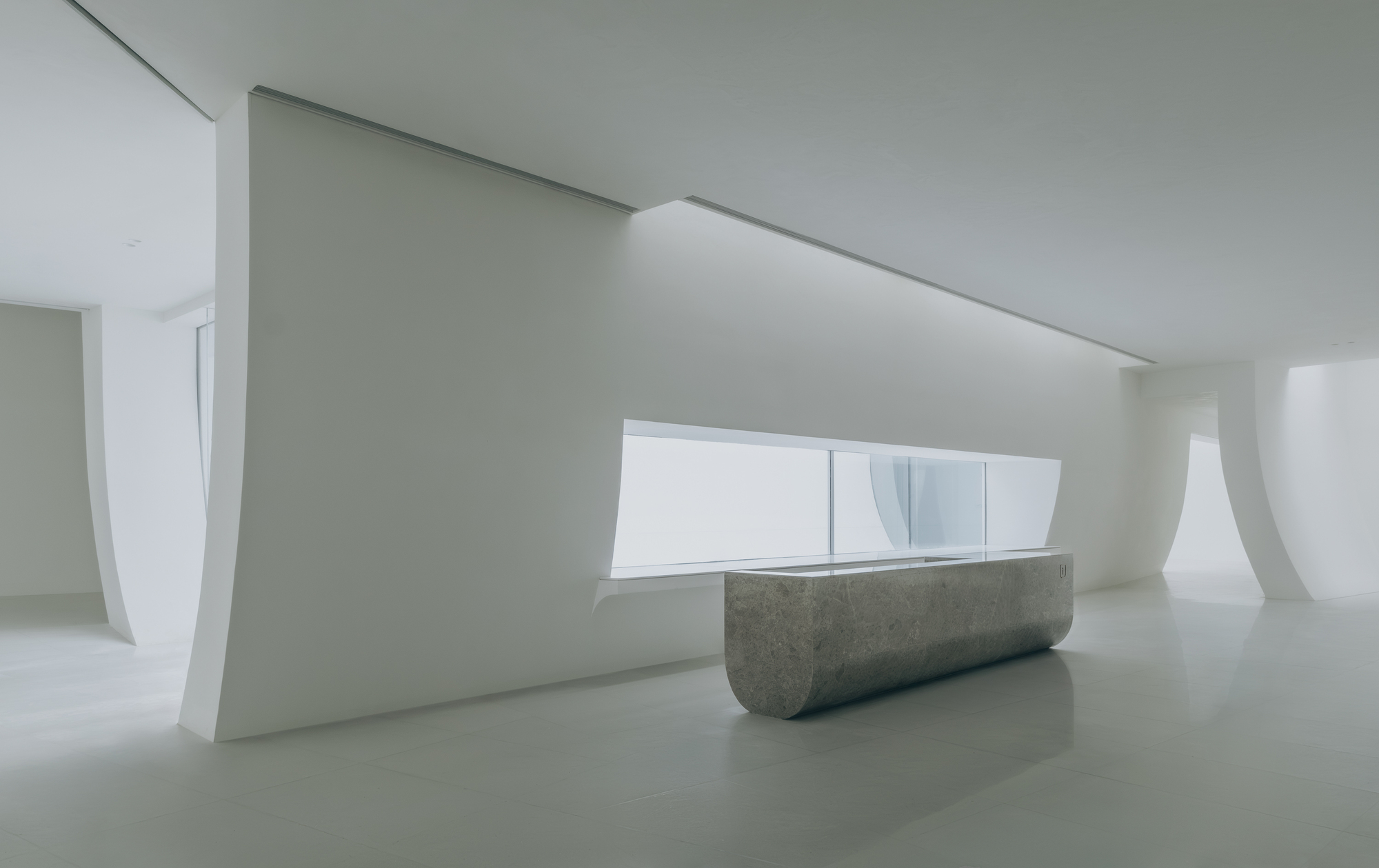 Reception desk in grey concrete beneath a horizontal window framed by curved white walls