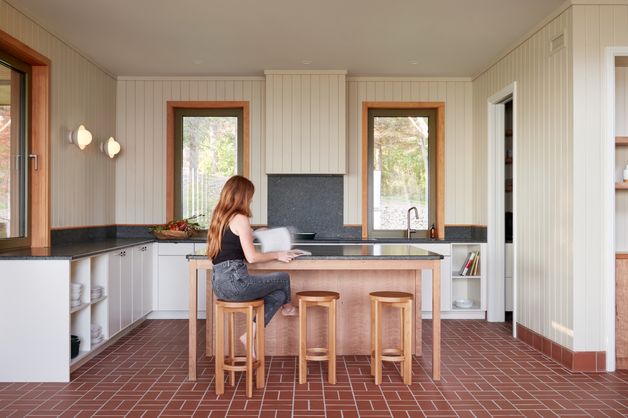Kitchen with timber island, three stools, and a person seated reading at the counter near two windows