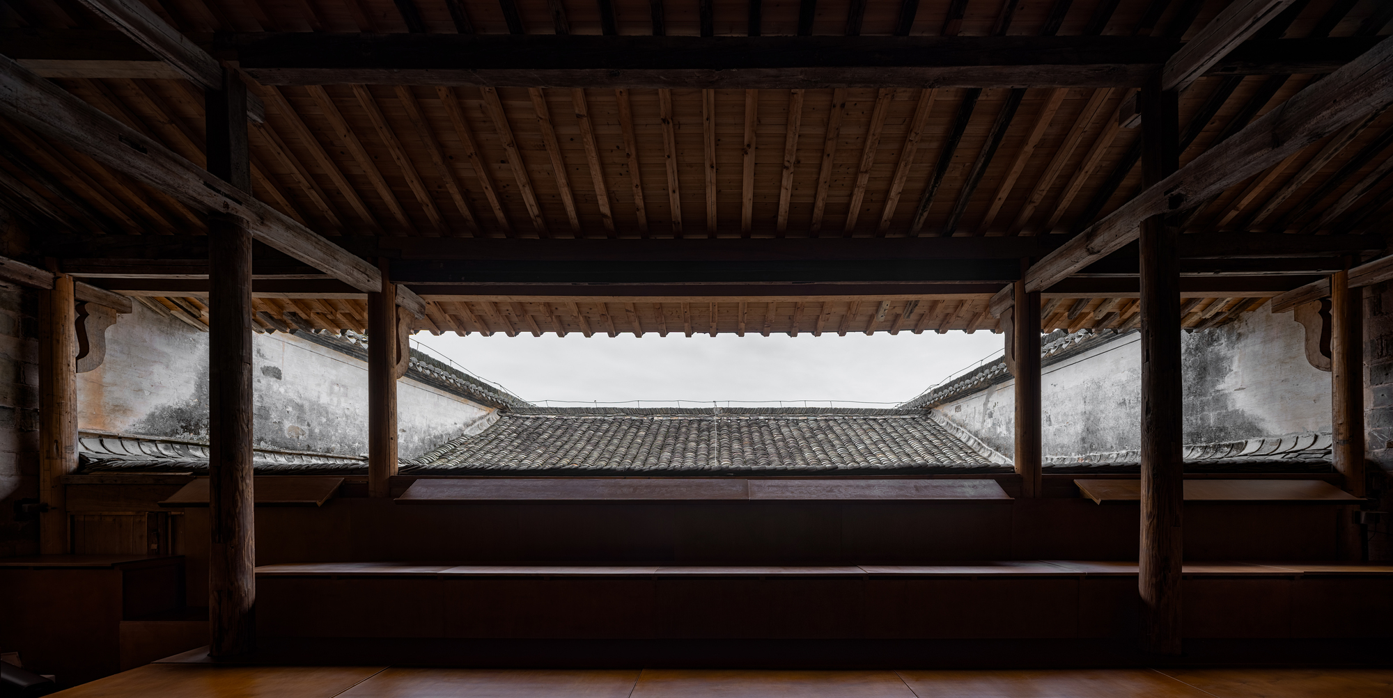 Framed view through timber structure to courtyard with tiled roof and white walls under overcast sky