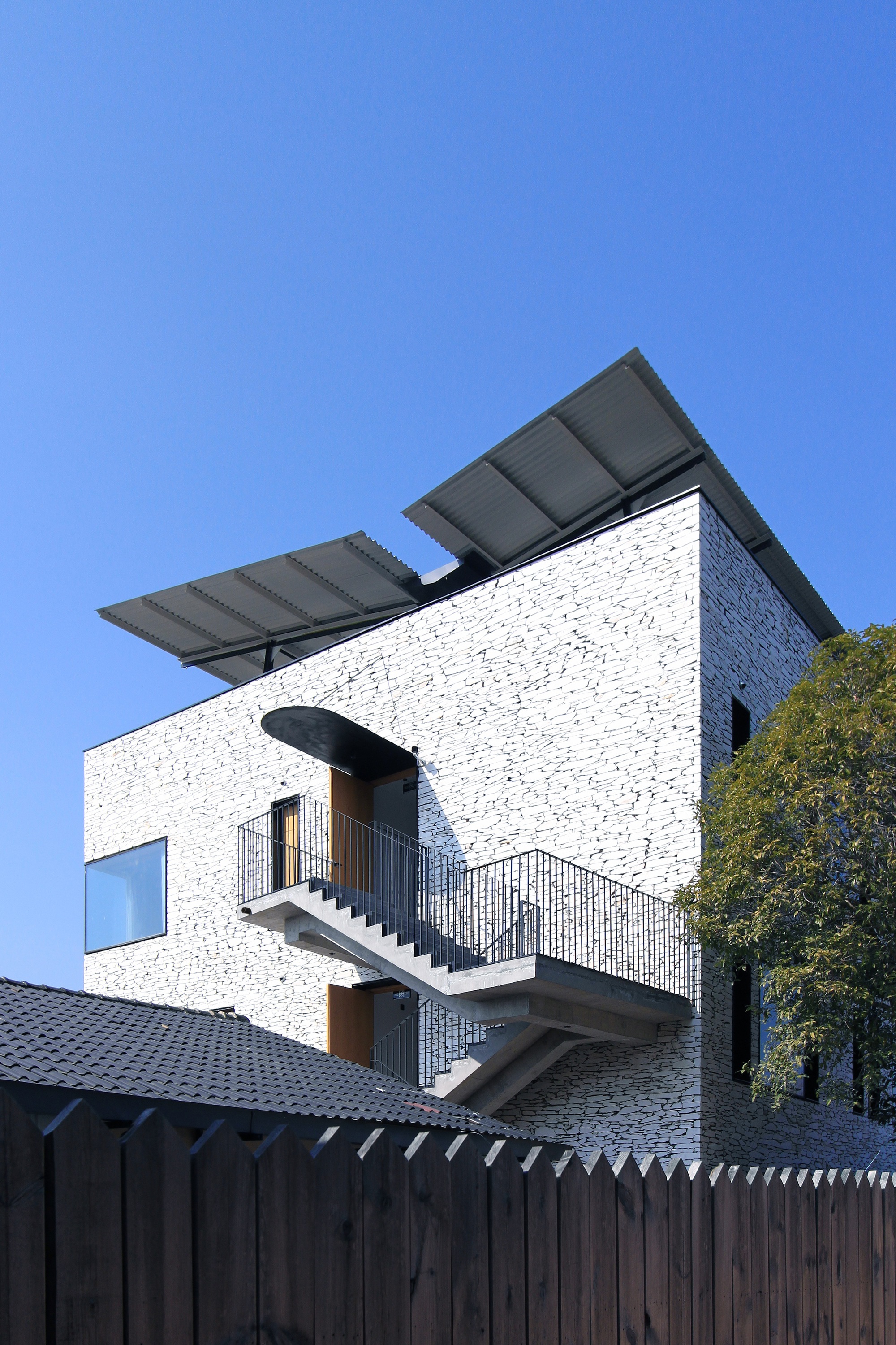 Textured stone facade with exterior stair leading to timber-framed balcony beneath tilted solar panel canopy