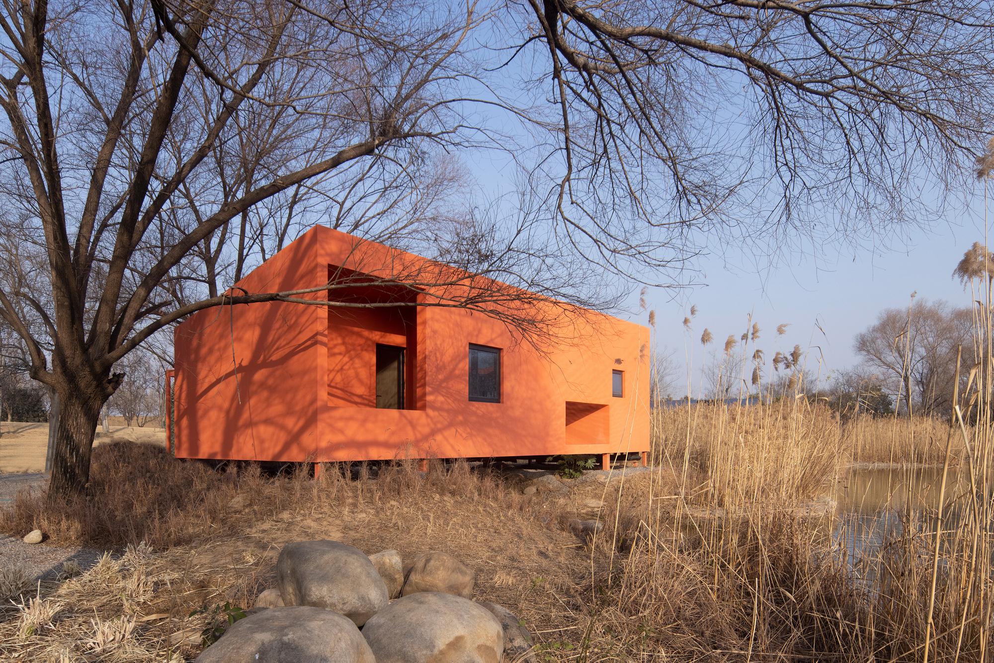 Red-orange facade with recessed openings on a raised platform among bare trees and dry vegetation