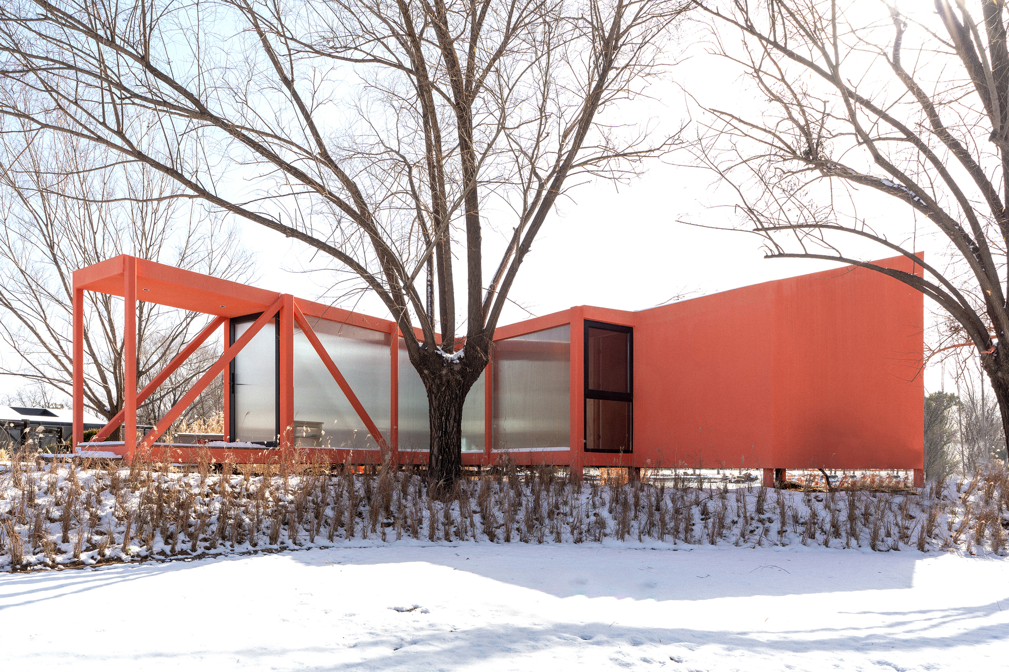 Coral-colored volume with steel-framed glazed enclosure and solid wall emerging from snow-covered ground