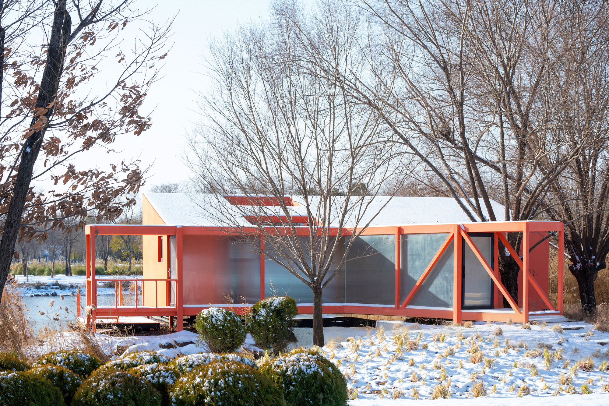 Red structural frame with glazed panels and white roof on snow-covered ground beneath bare trees