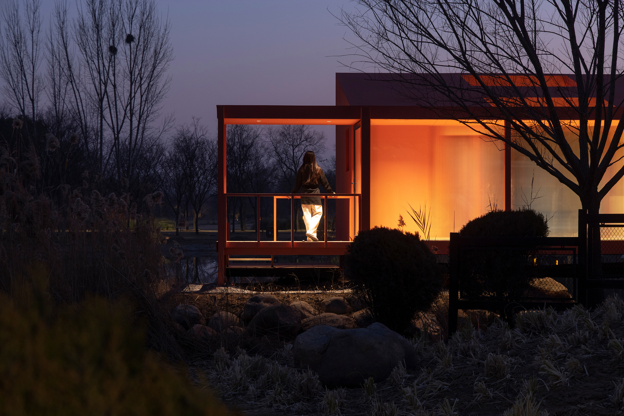 Exterior deck with figure standing at twilight as warm light spills from the glazed interior
