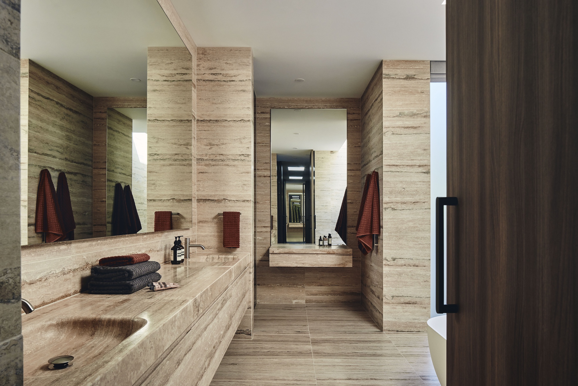 Bathroom vanity lined in travertine with mirrored wall reflecting the corridor beyond