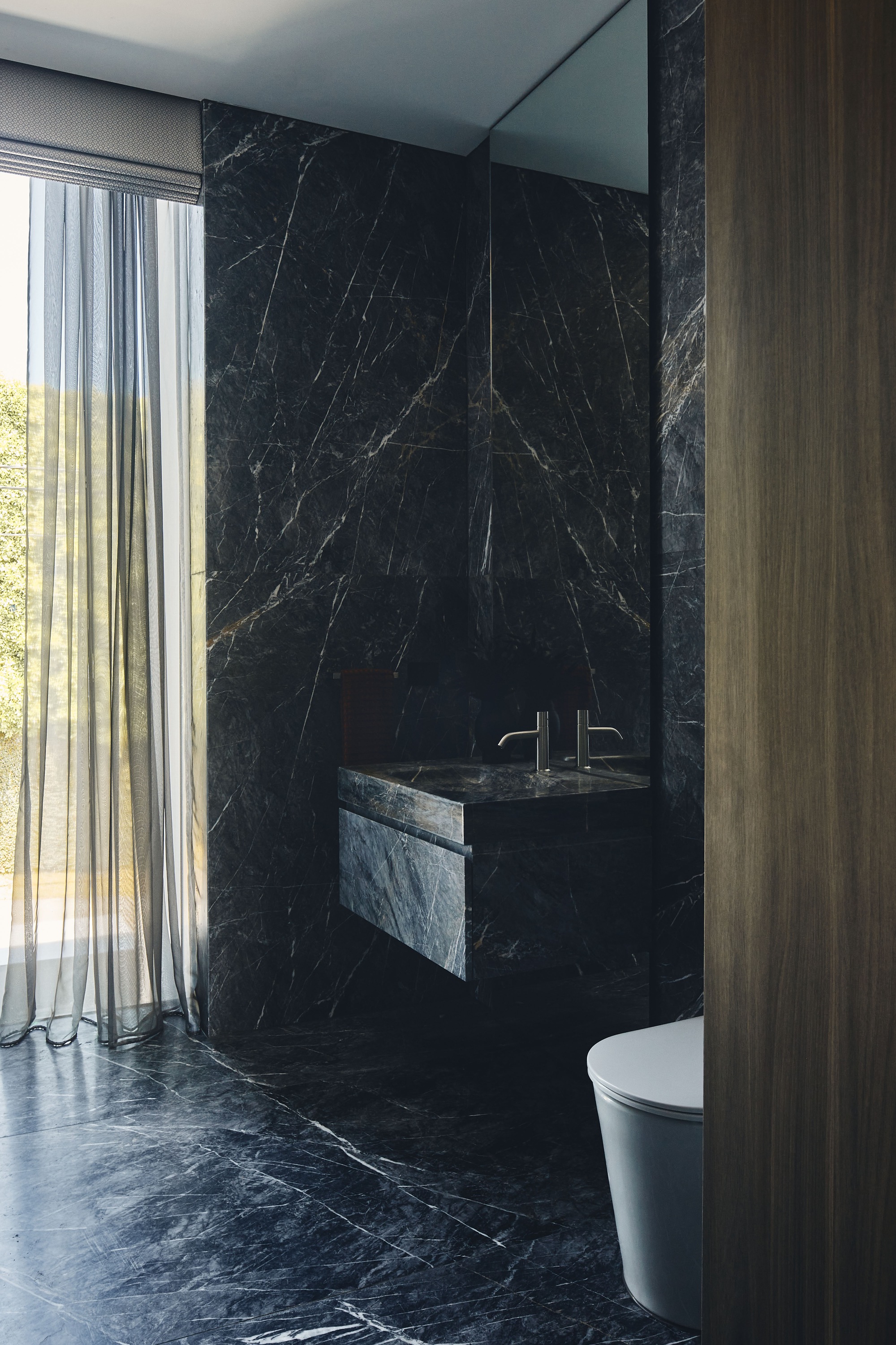 Bathroom with black marble walls, floating sink, and sheer curtains revealing trees outside