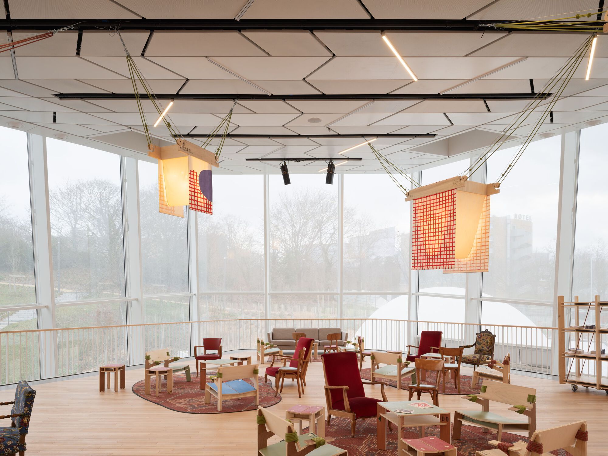Glass-walled upper floor activity room with geometric ceiling, suspended fabric screens, and misty trees beyond