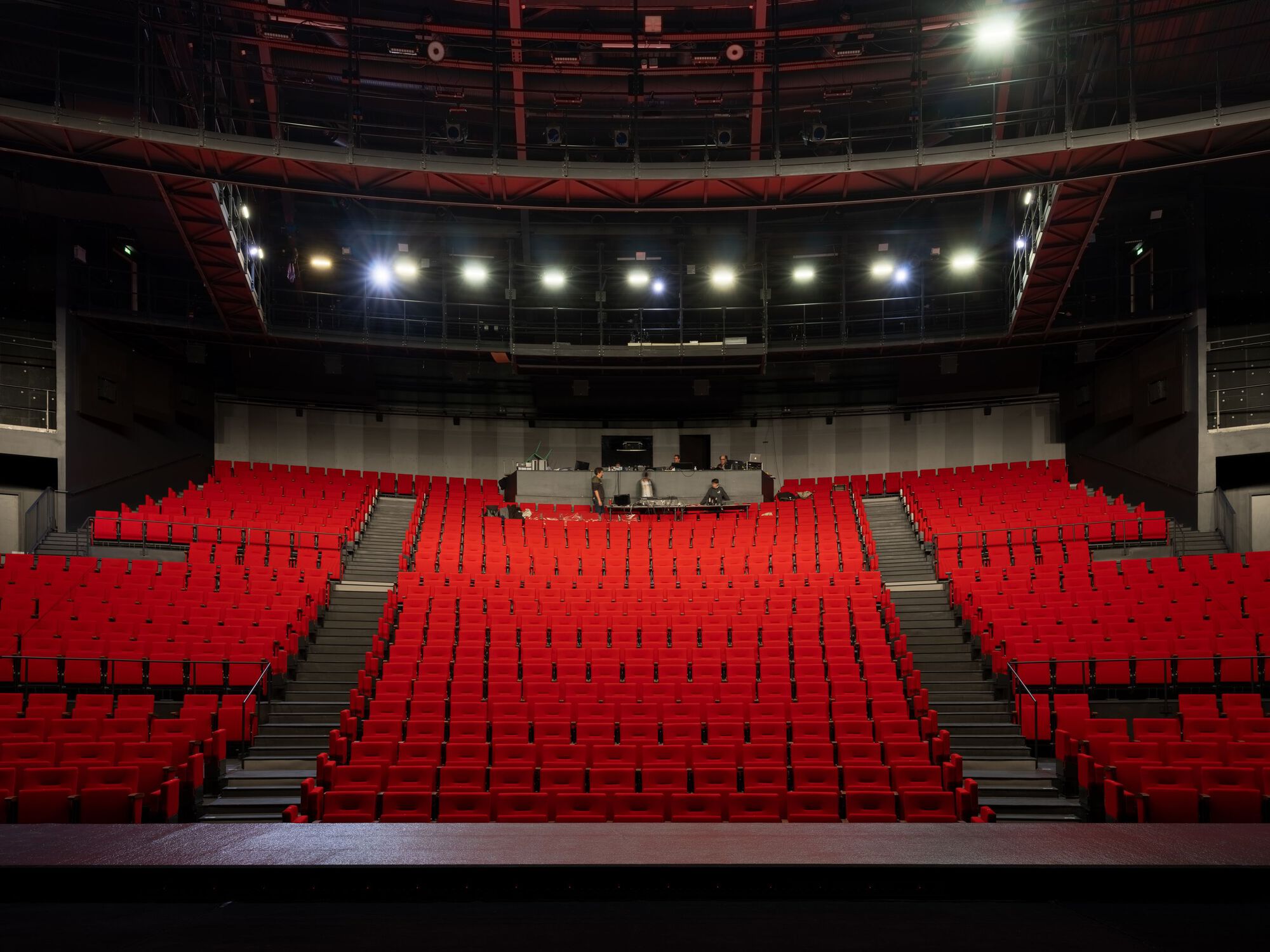 Theater auditorium with tiered red seating, exposed steel trusses, and stage-level lighting