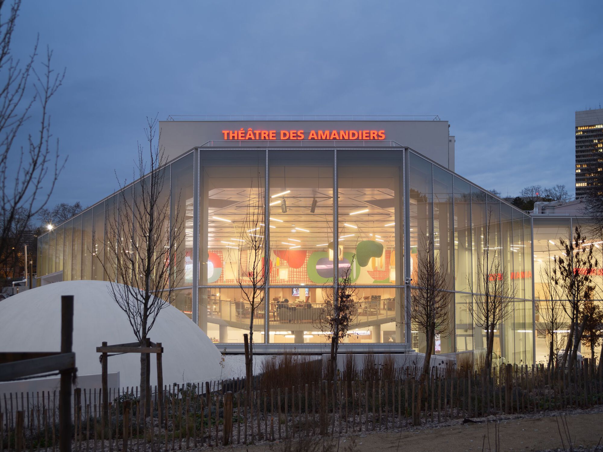 Glass facade of the theatre building illuminated at dusk with bare winter trees in the foreground