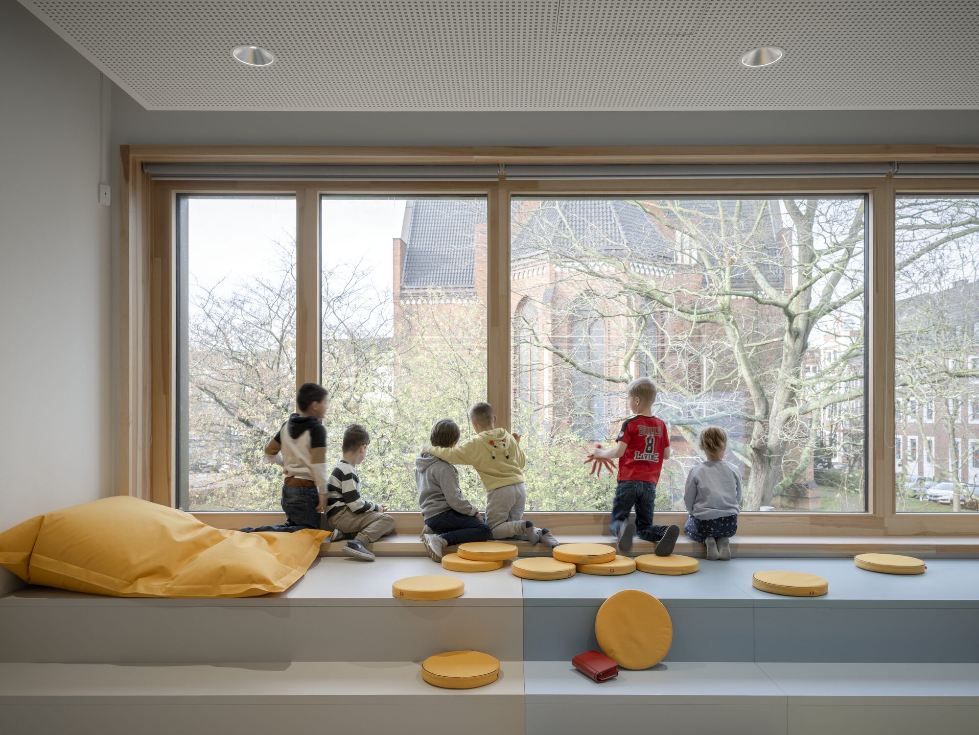 Window bench with children seated facing bare branches and brick buildings outside
